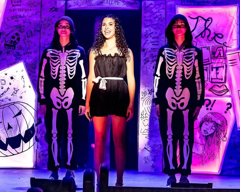 A woman standing between two people dressed in skeleton costumes, with colorful Halloween-themed background decorations, including drawings of a pumpkin and a girl with curly hair.