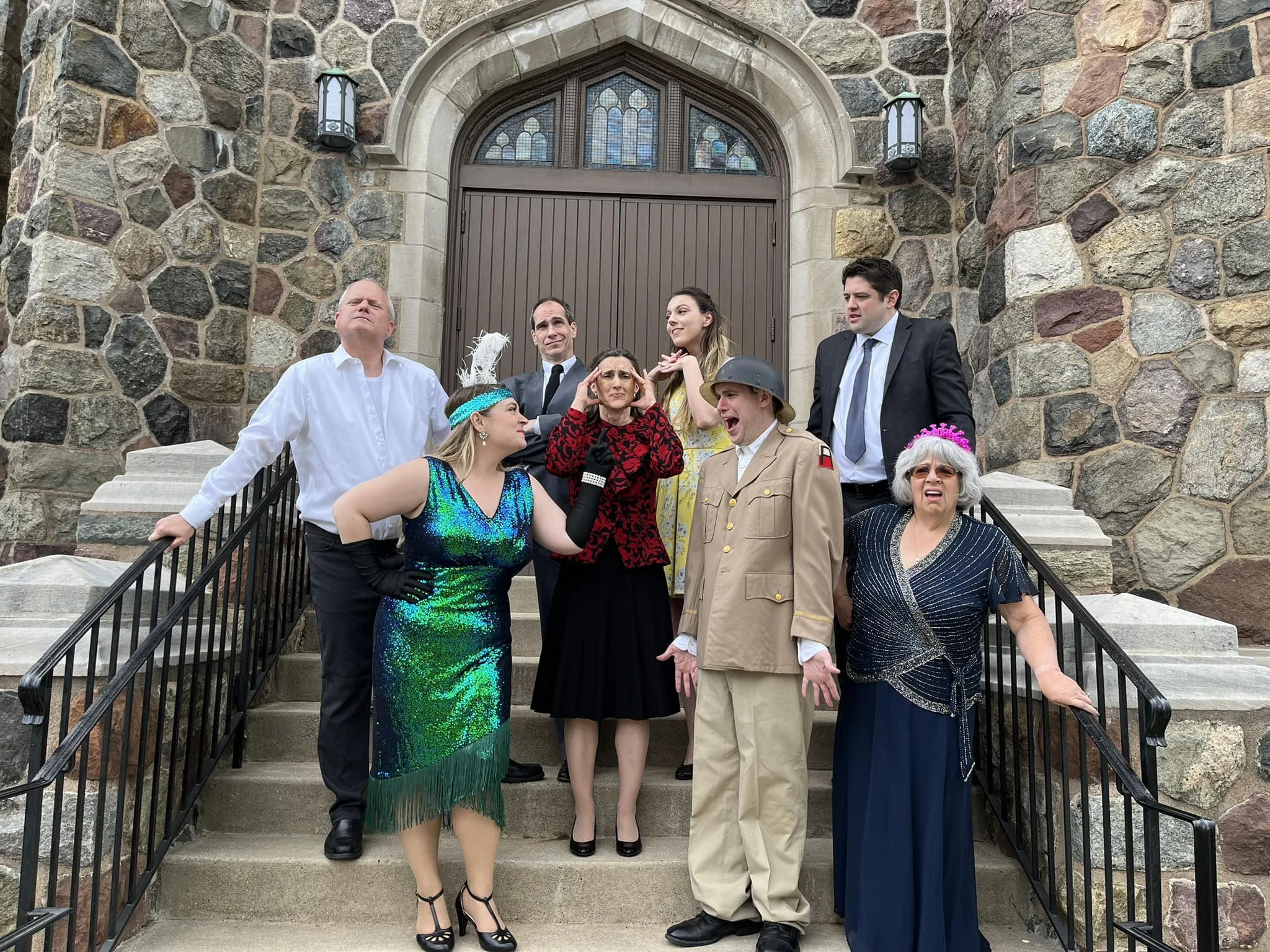 A group of nine people dressed in costumes for a play or event standing on the steps of a stone building. The costumes vary from formal suits, vintage attire, military uniform, and glamorous dresses, with some individuals making exaggerated facial ex