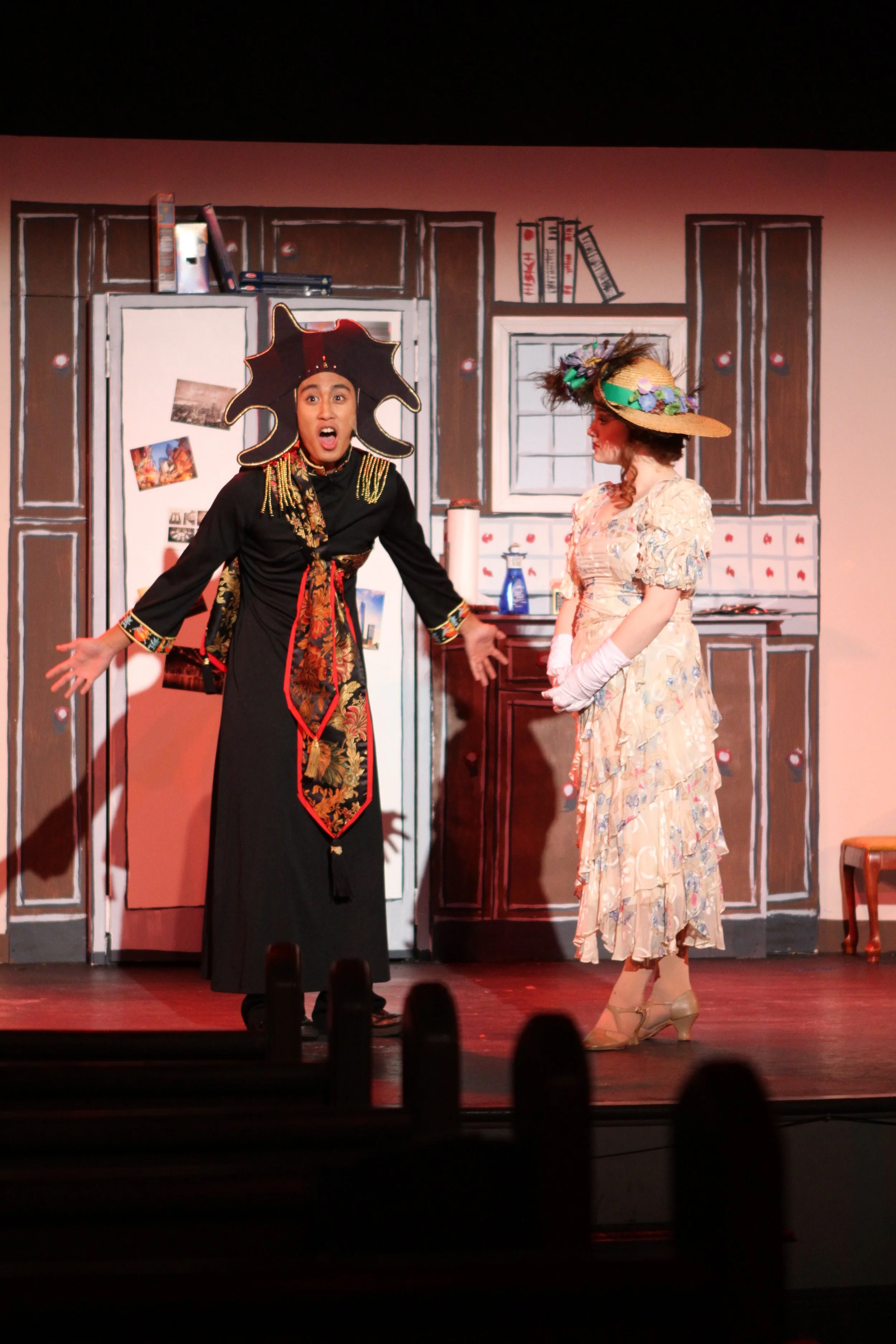 Two women performing on a stage with a kitchen set background, one in an elaborate black costume with a large hat and the other in a floral dress with a wide-brim hat.