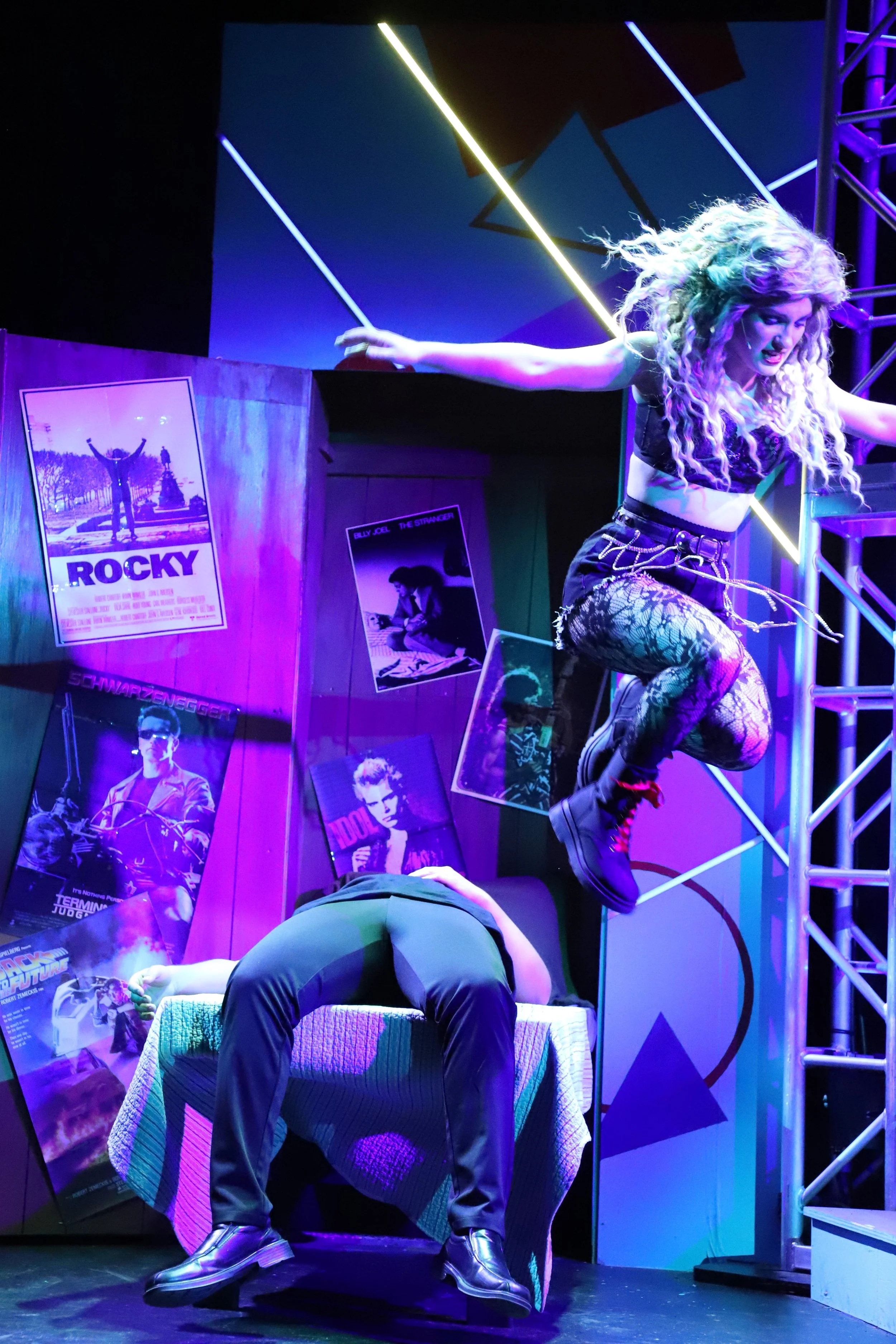 A woman jumping off a table in a theatrical performance, with a man bent over the table. The background features colorful posters of movies like 'Rocky' and others, with neon lights and stage equipment.
