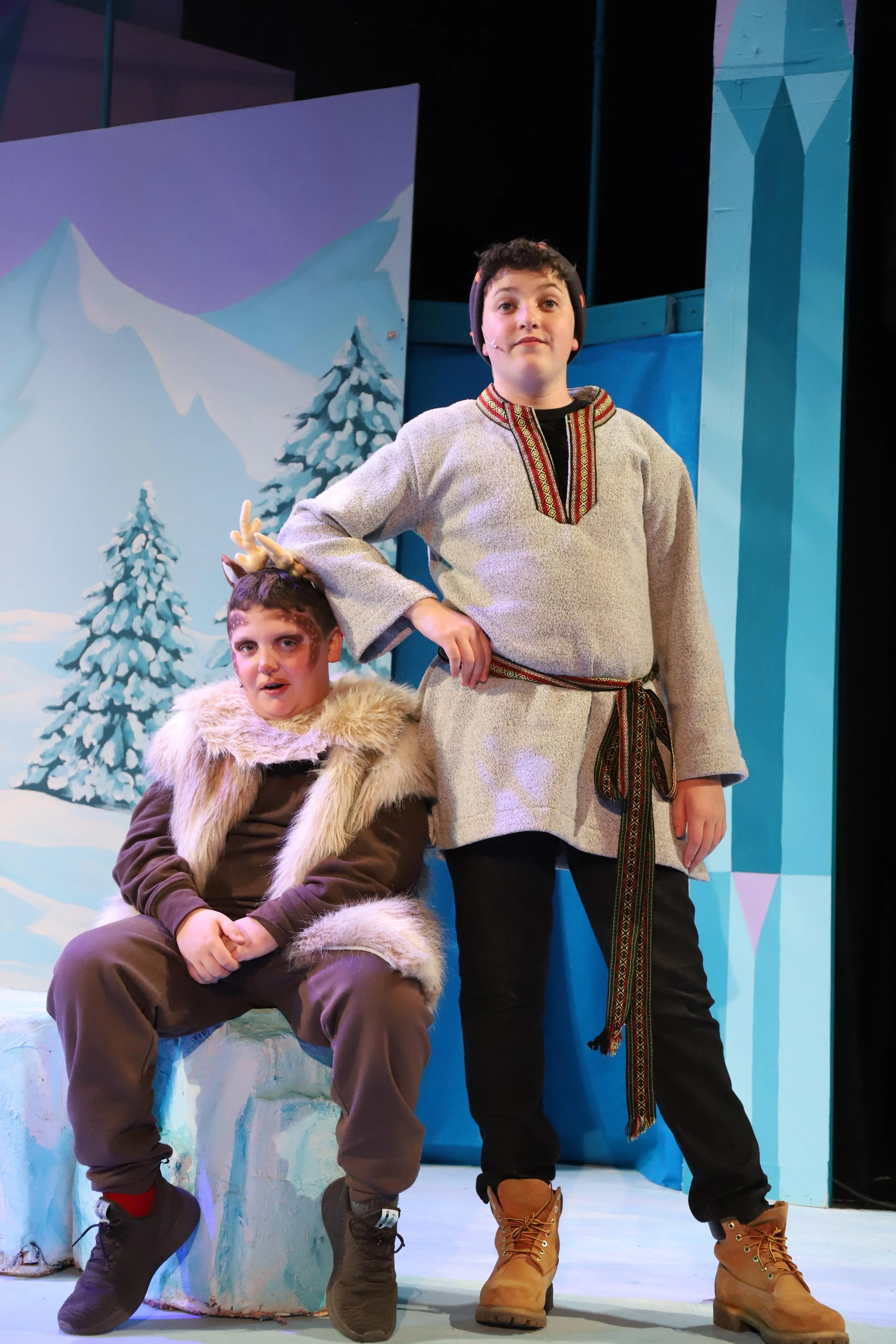 Two young boys in costume performing on a winter-themed stage, one seated with reindeer antlers and faux fur clothing, the other standing in a traditional tunic and boots against a backdrop of snow-covered trees.