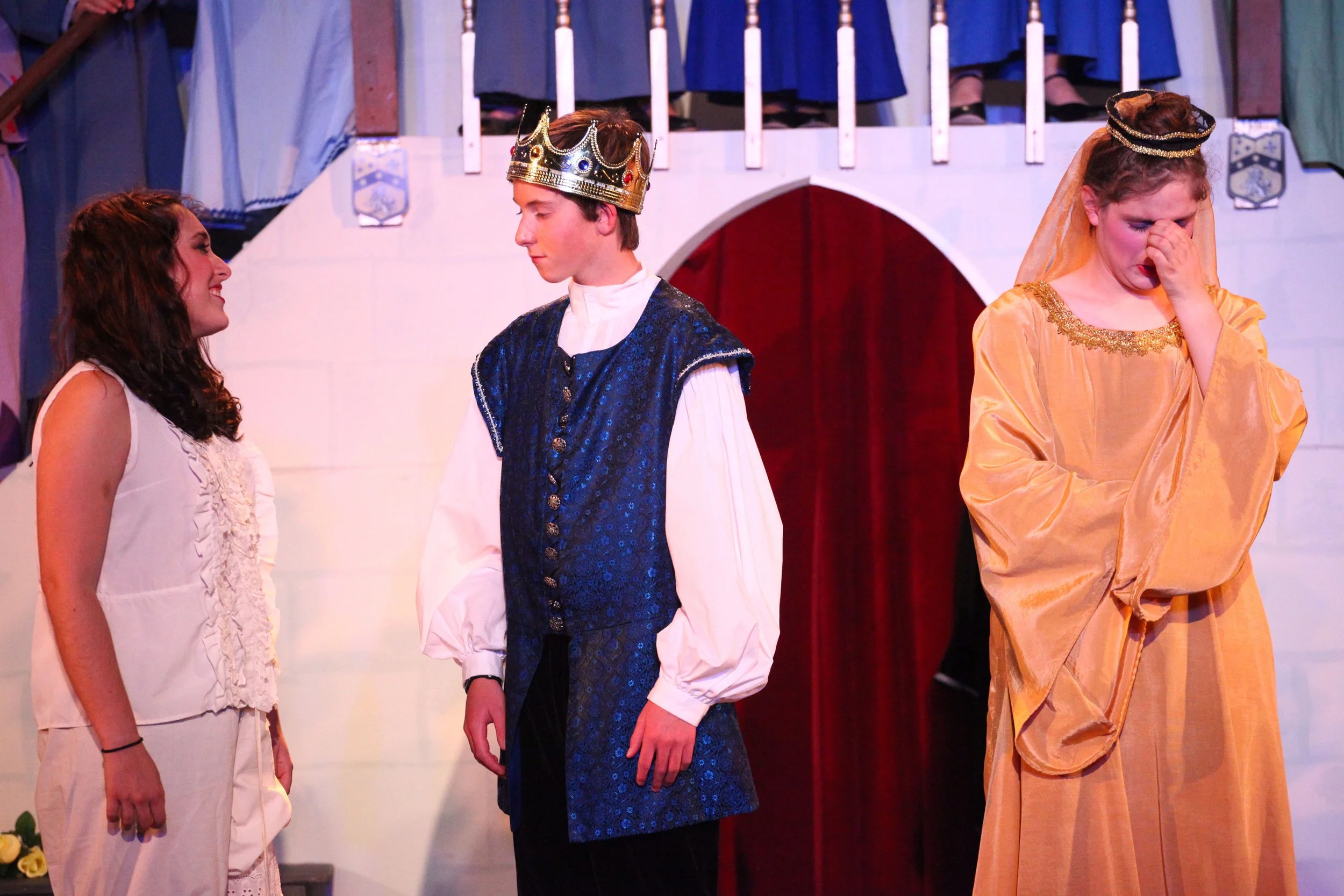 3 actors in Once Upon a Mattress, in front of a castle; the woman on the left is wearing a white dress, the man in the middle is dressed as a king in a blue robe and crown, and the woman on the right is wearing a gold dress and a crown, looking upset