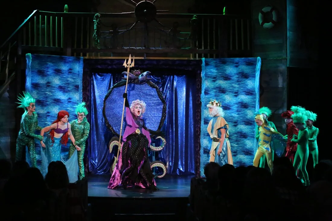 Scene from a theatrical performance of The Little Mermaid with actors dressed as mermaids and sea characters on stage with underwater-themed backdrops.