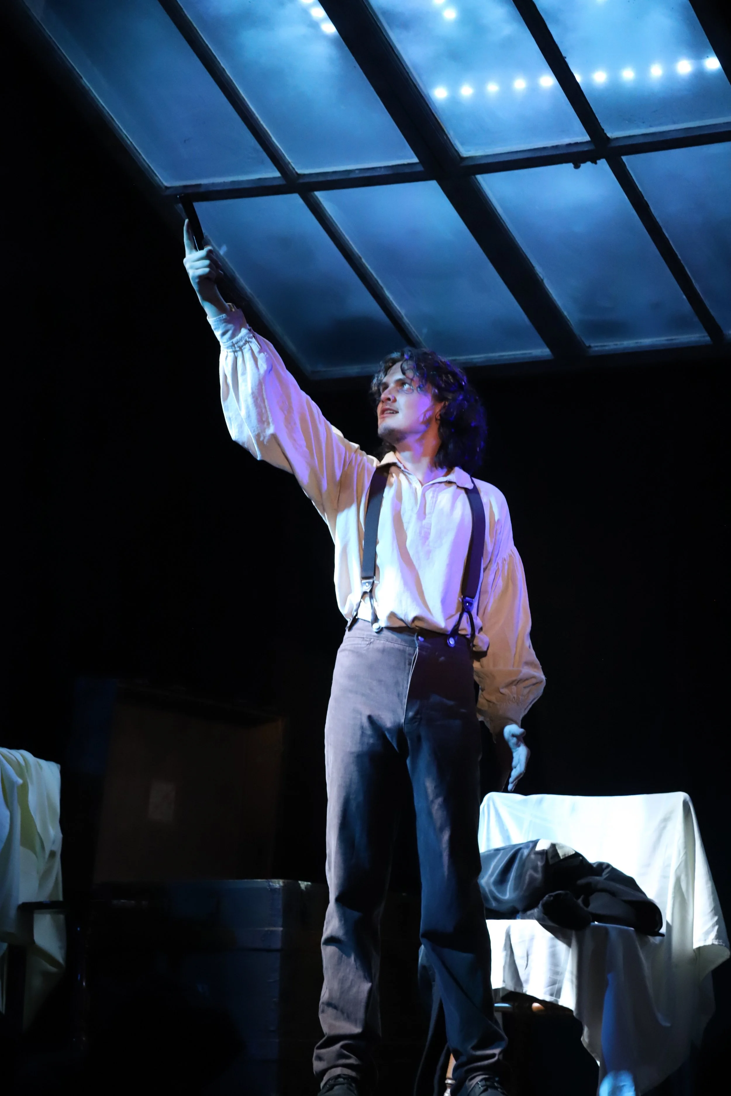 A person dressed in historical attire, wearing suspenders and a loose shirt, stands on stage under dramatic lighting. They are reaching up toward a large window or skylight on the ceiling, with a contemplative expression.