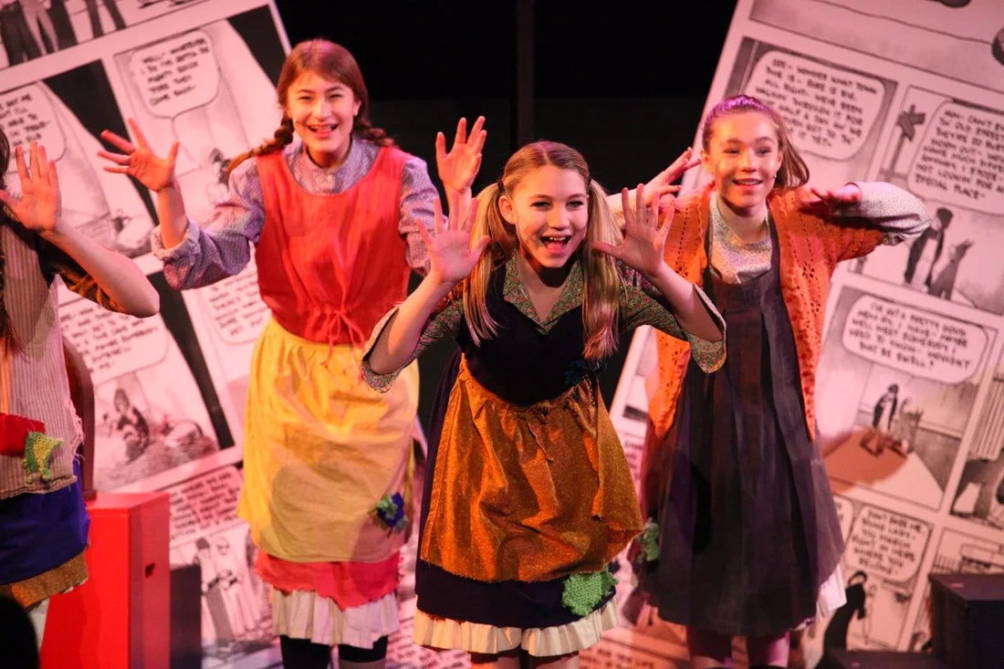 Three young girls on stage, smiling and waving, in costume for a theatrical performance with comic strip background panels.