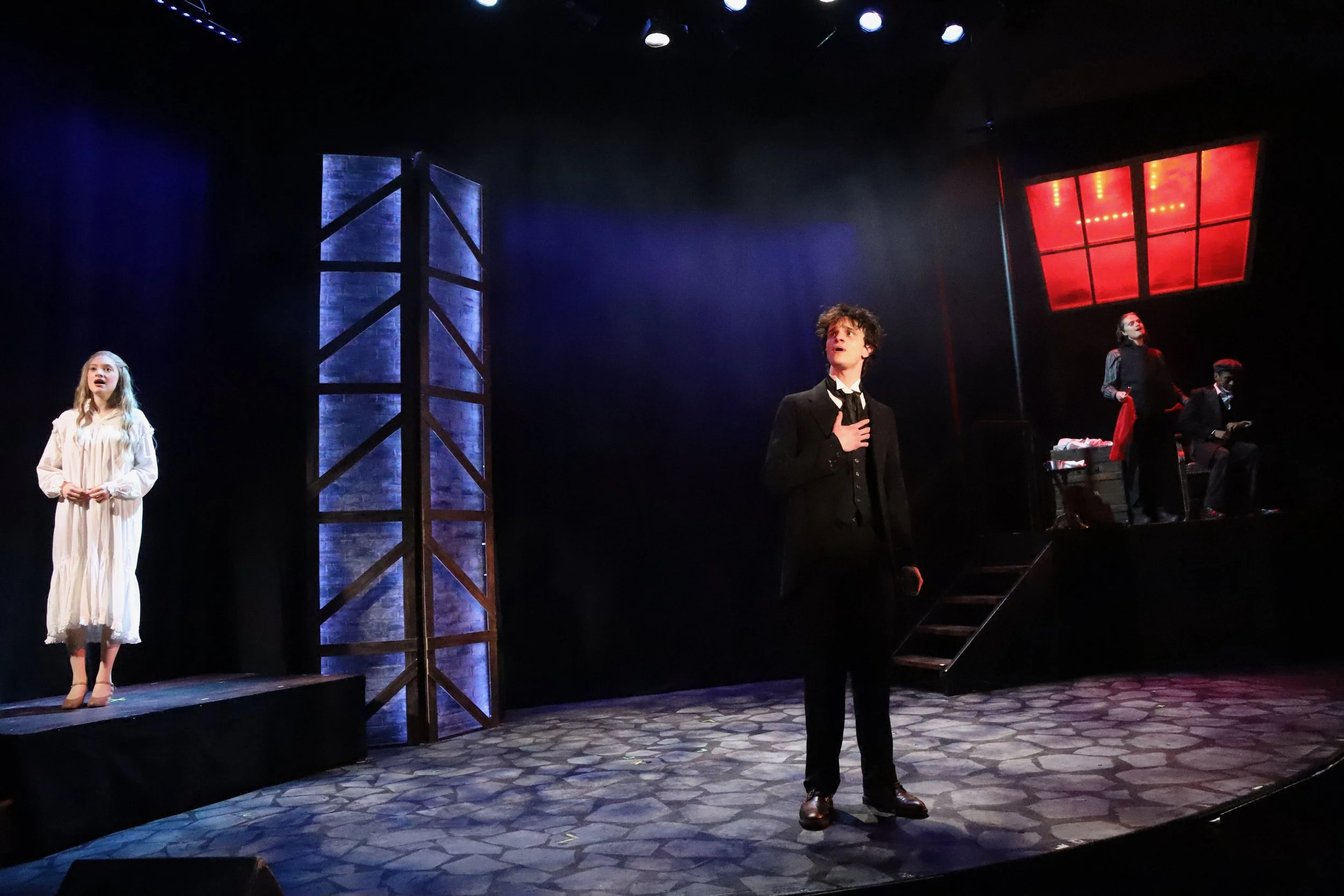 Actors performing on stage with a woman in a white dress on the left, a man in a black suit singing in the center, and two actors sitting and standing on the right, with blue and red lighting effects.