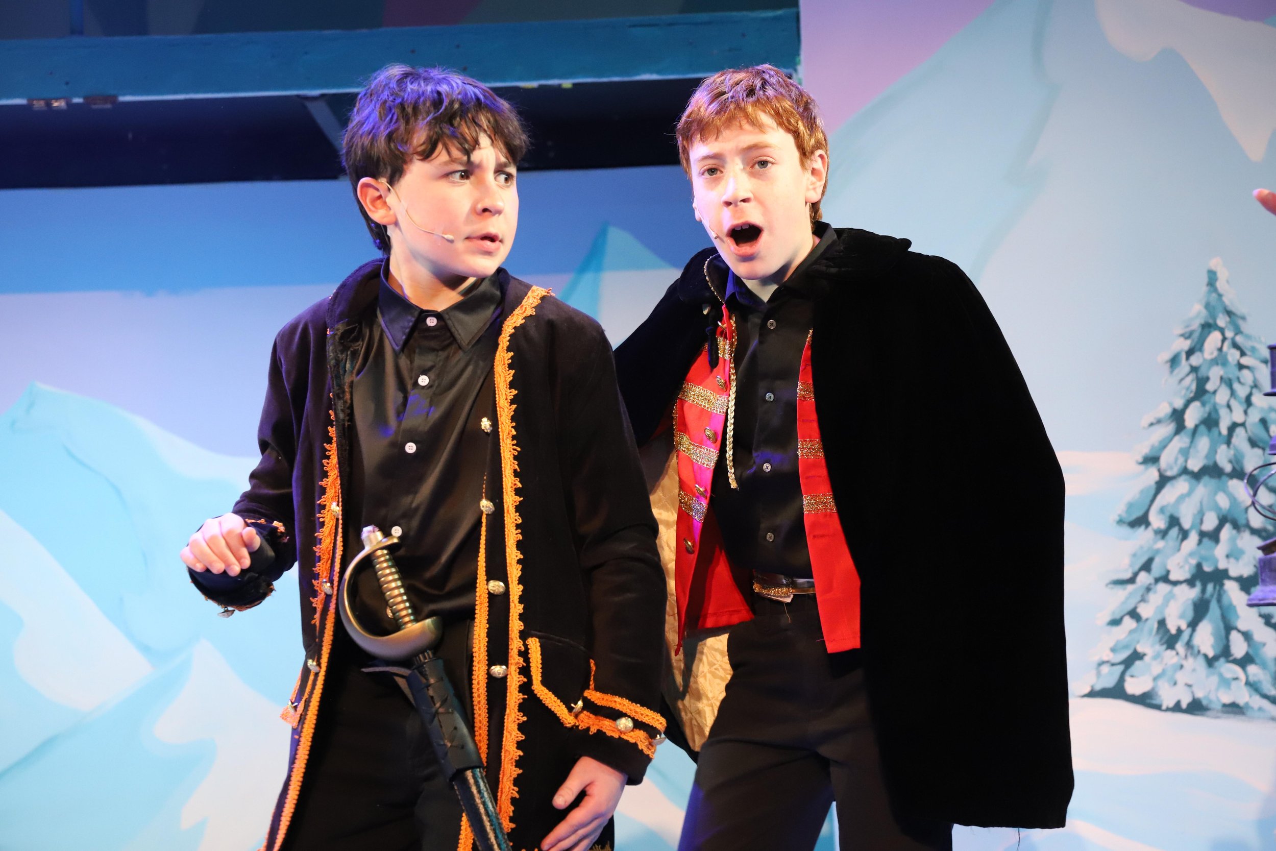 Two young boys performing on stage with snowy mountain and pine tree backdrop, dressed in costumes, engaged in a theatrical scene.
