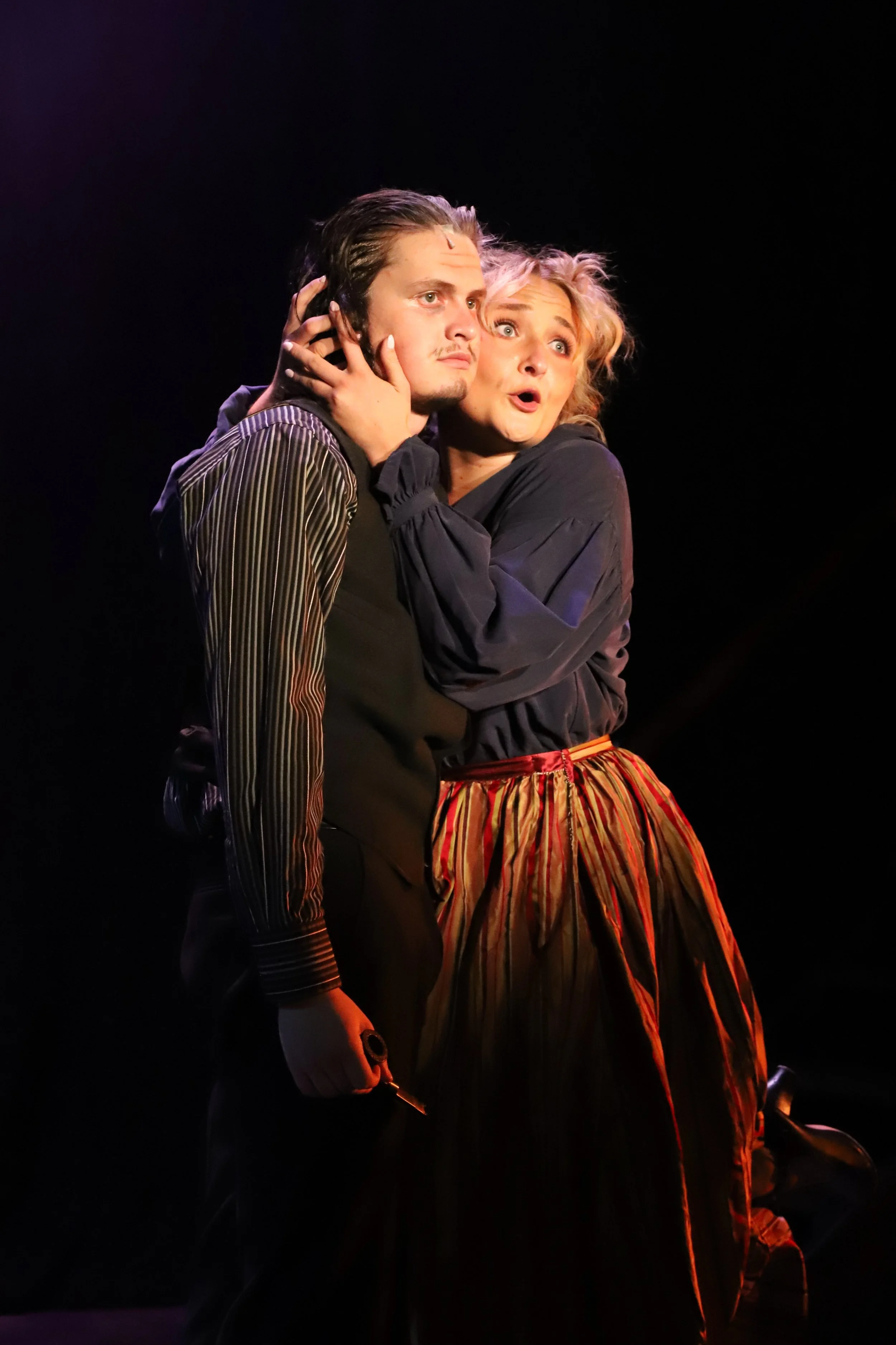 Two actors perform on stage, a man with dark hair and a woman with curly blonde hair, in a dramatic scene with a black background.
