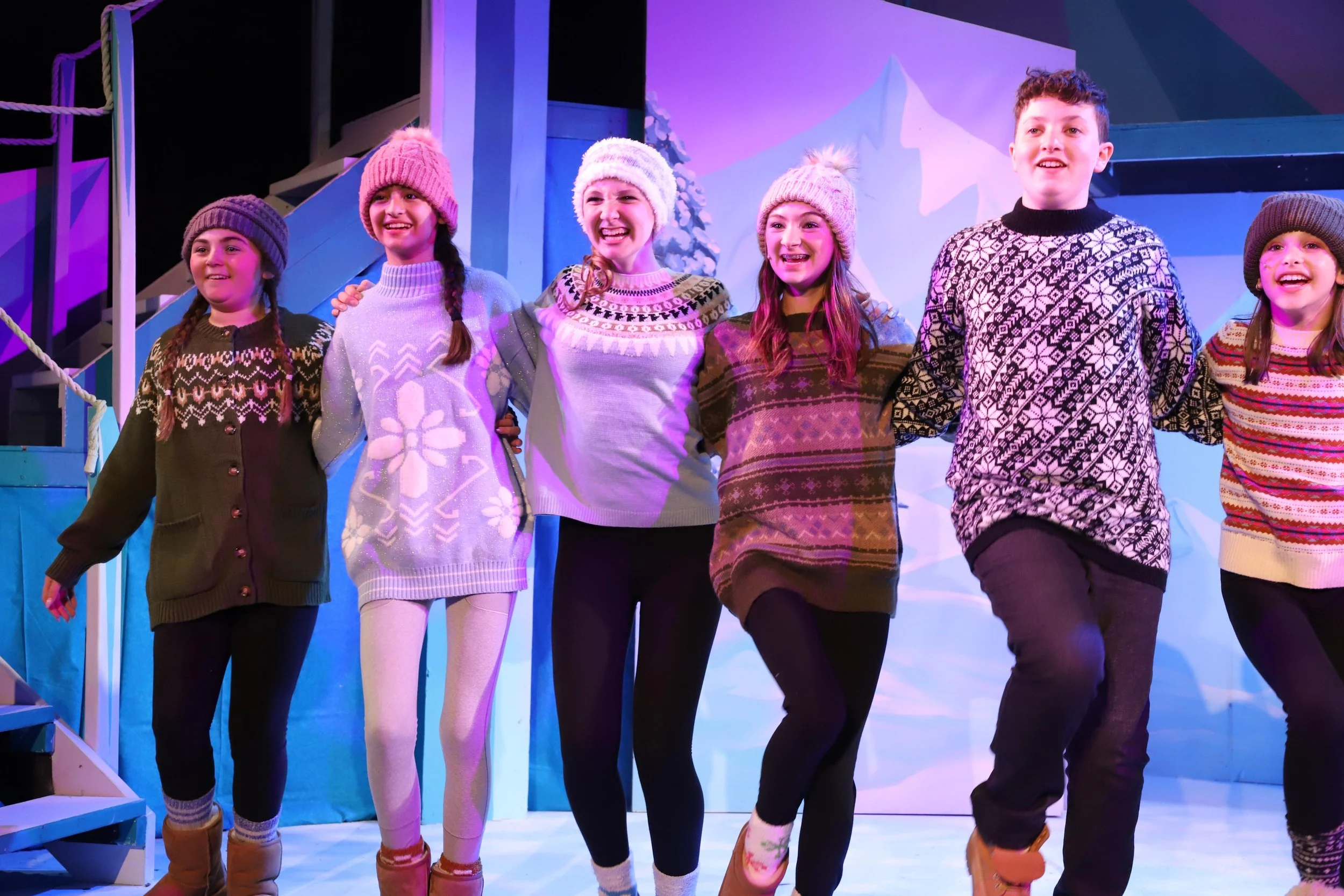 A group of seven children, dressed in winter sweaters, hats, and boots, holding hands and smiling on a stage with a winter-themed backdrop.