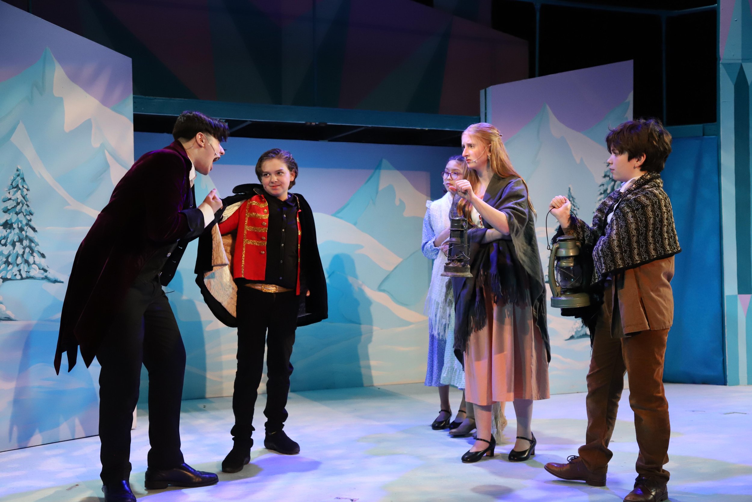 Six actors performing a scene on stage with a winter mountain backdrop, dressed in costumes from Frozen Jr with some holding lanterns.