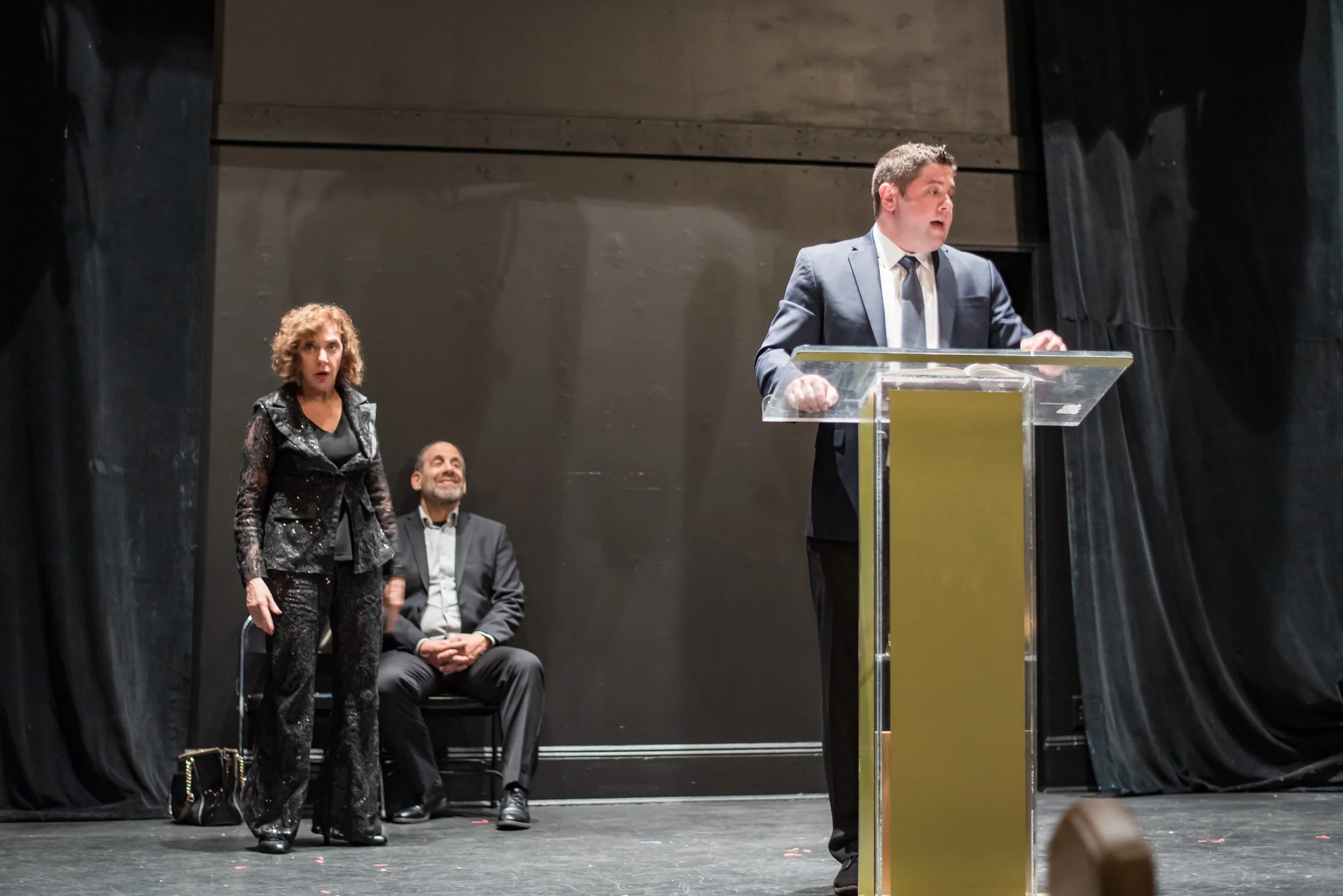 Three actors on stage, two seated and one standing at a clear podium, performing in a theatrical production of Grandma Sylvia's Funeral with black curtains and plain background.