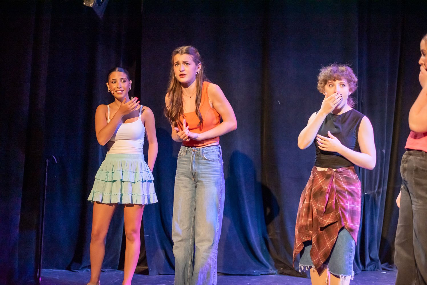 Four young women on stage during a theatrical performance, with black curtains in the background. One woman in a white top and tiered skirt looks surprised or concerned, another in an orange tank top and jeans appears to be explaining or reacting, th