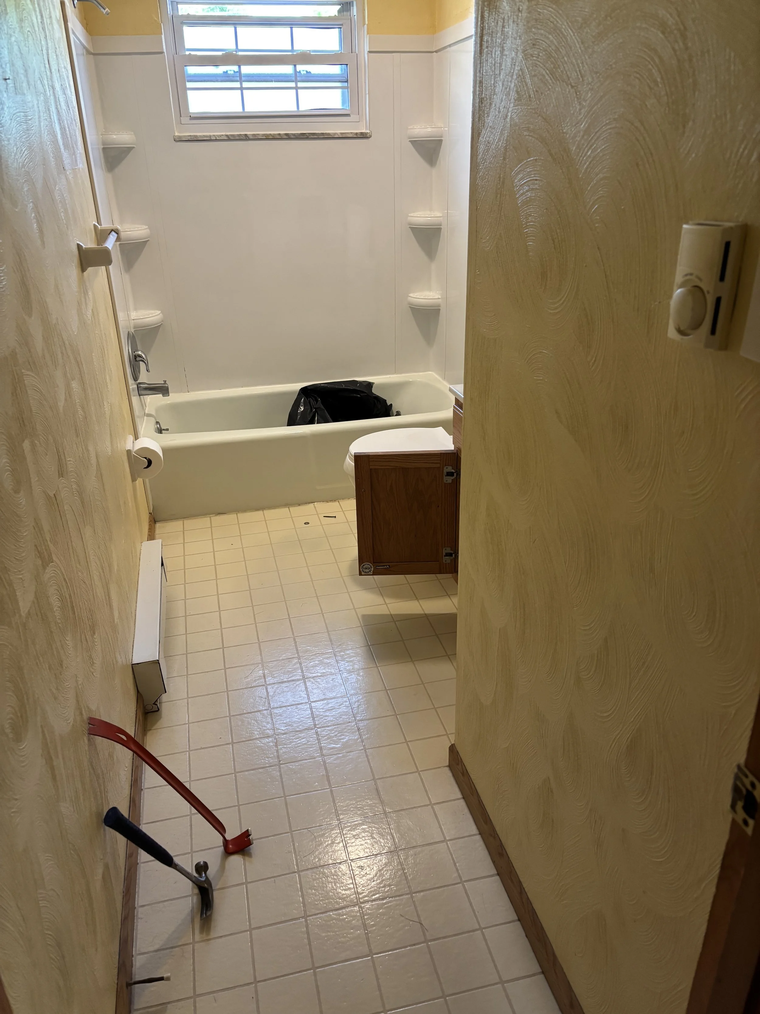 A small bathroom with yellow painted walls, a bathtub with white wall surrounding it, a small window above, a wooden cabinet near the toilet, and a floor with beige tiles. There are cleaning tools on the floor.