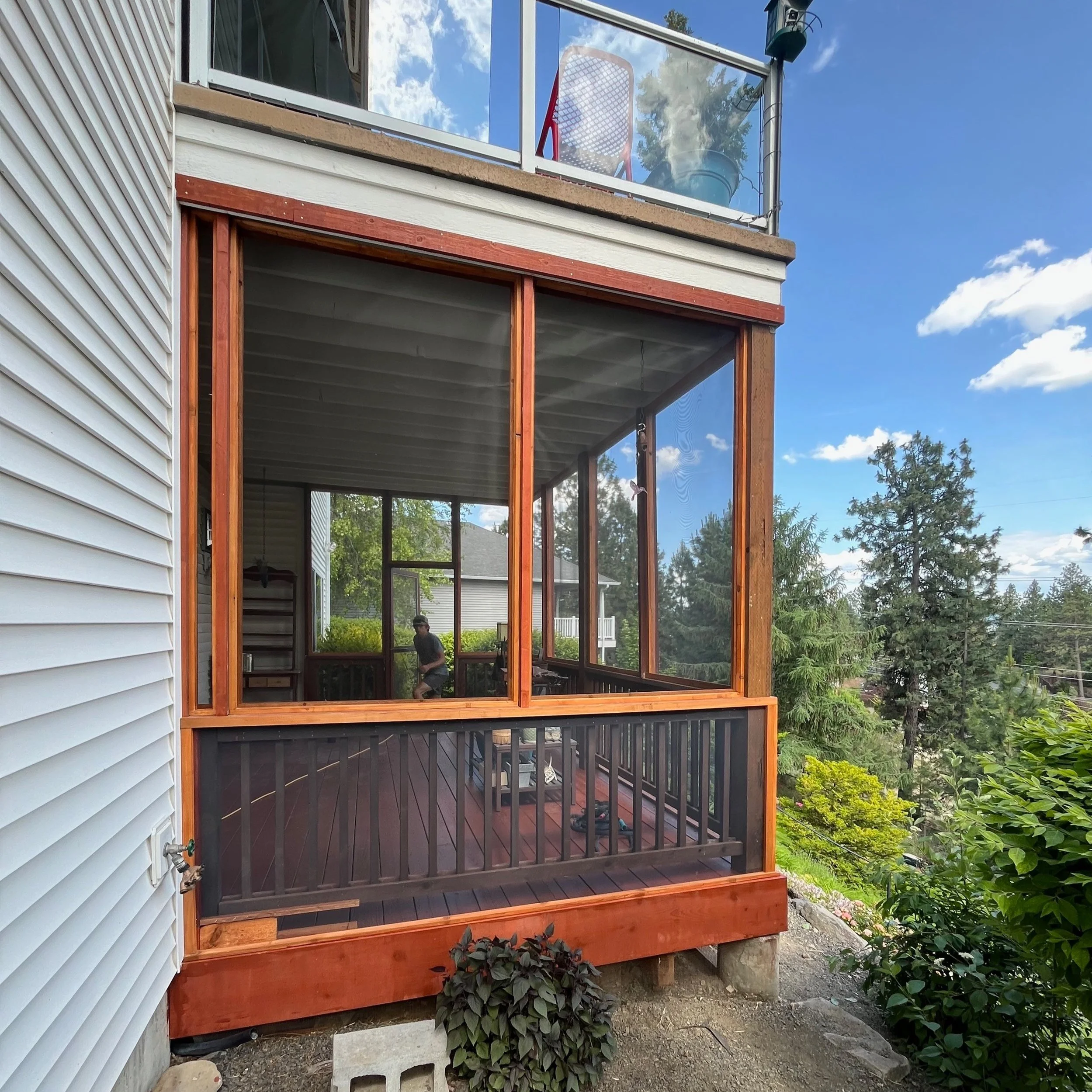 A screened-in porch attached to a two-story house, with a person working inside. The porch has wooden framing, black railing, and is surrounded by greenery and trees under a blue sky.