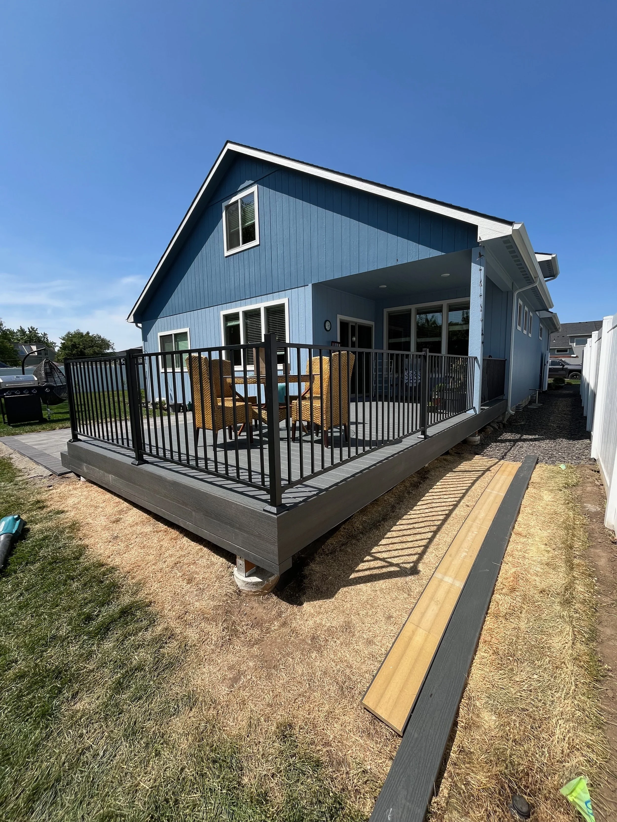 Backyard view of a blue house with a elevated wooden deck, black railing, patio furniture, and a sliding glass door on a sunny day.