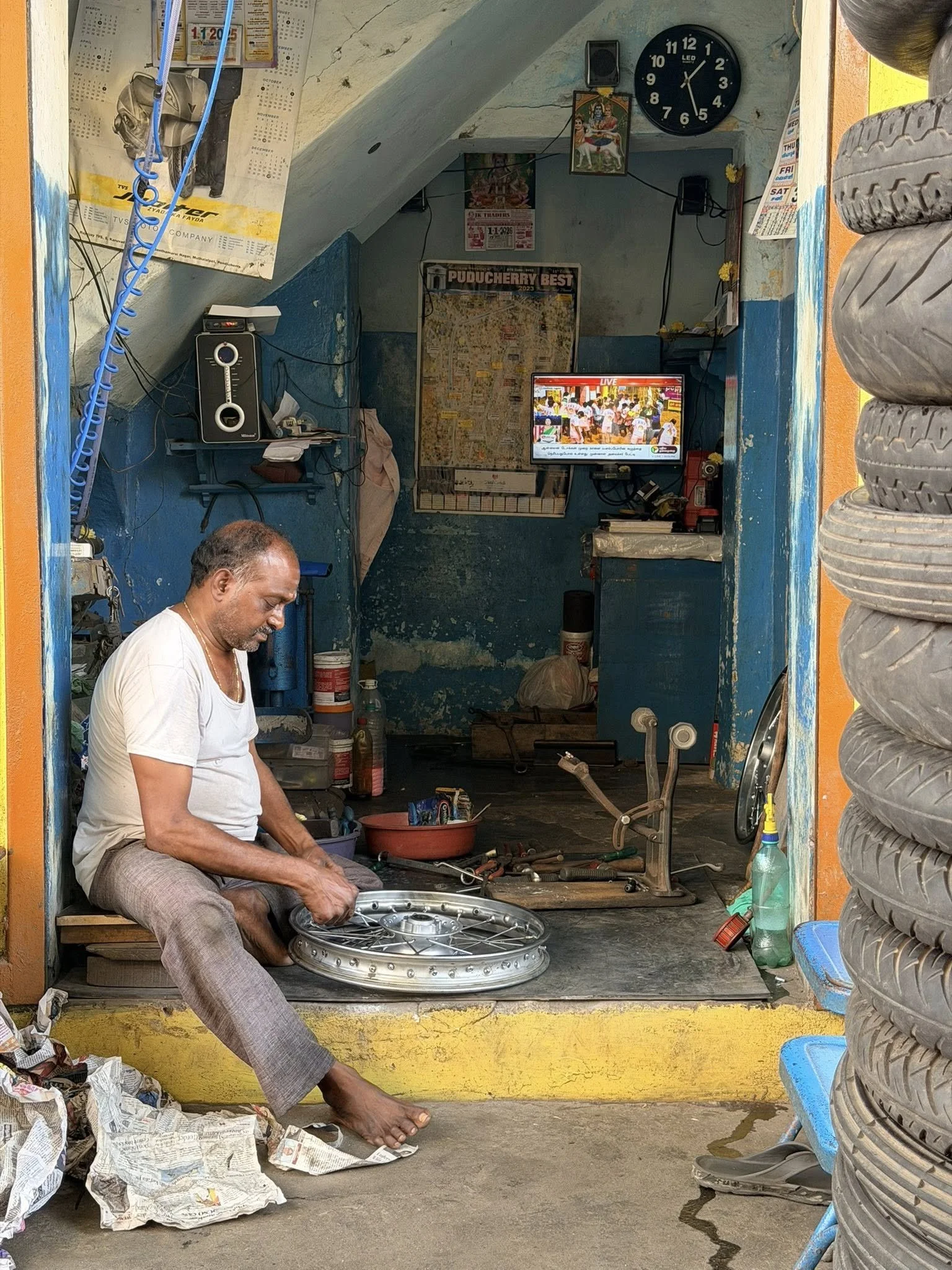 A man sitting on the floor of a tire shop, working on a metal wheel rim. The shop has various tools, tires, a wall clock, and a small television screen displaying a live broadcast. The environment appears to be a small, local auto repair shop with wo