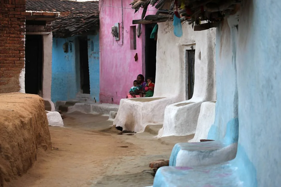A dirt street in a rural village with colorful adobe houses painted pink, blue, and white. Several women and children sit outside on the steps of one house.