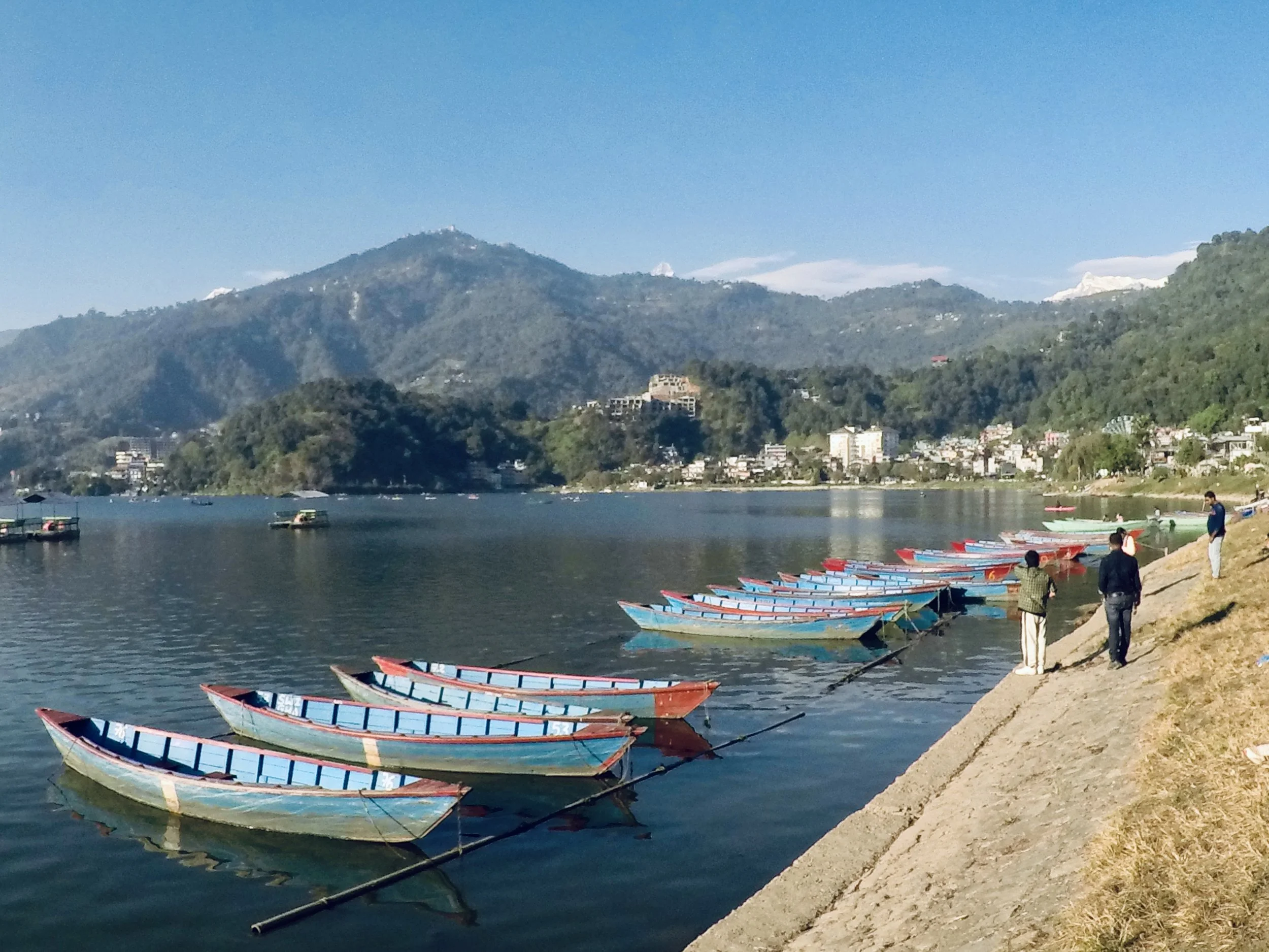 A lakeside scene with multiple colorful rowboats tied along the shore, a few people walking on the bank, and a backdrop of green mountains under a clear blue sky.