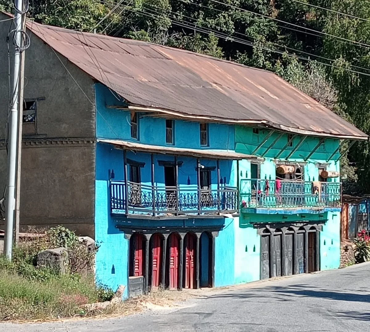 A two-story house with a rusty metal roof, blue walls, and balconies with black wrought iron railings; the house is next to a road in a rural area.