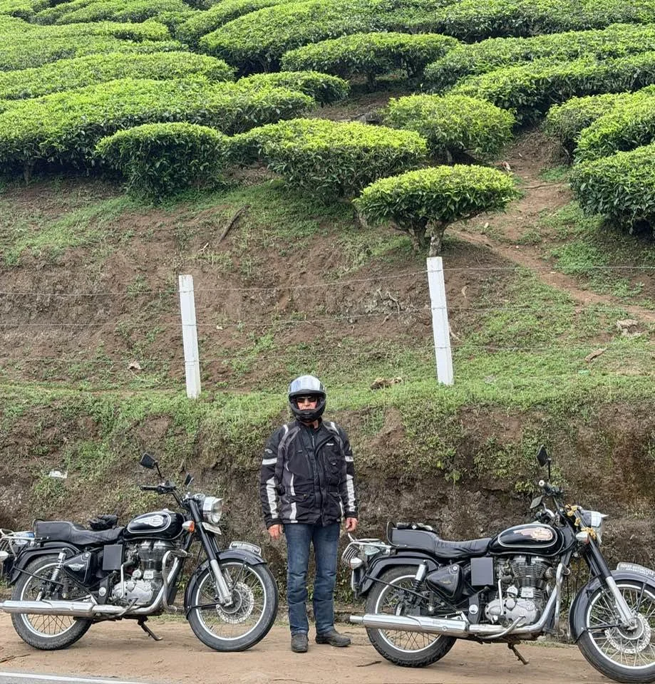 A man in motorcycle gear and a helmet stands between two black cruiser motorcycles on a dirt road, with a hillside covered in green bushes and trees behind him.