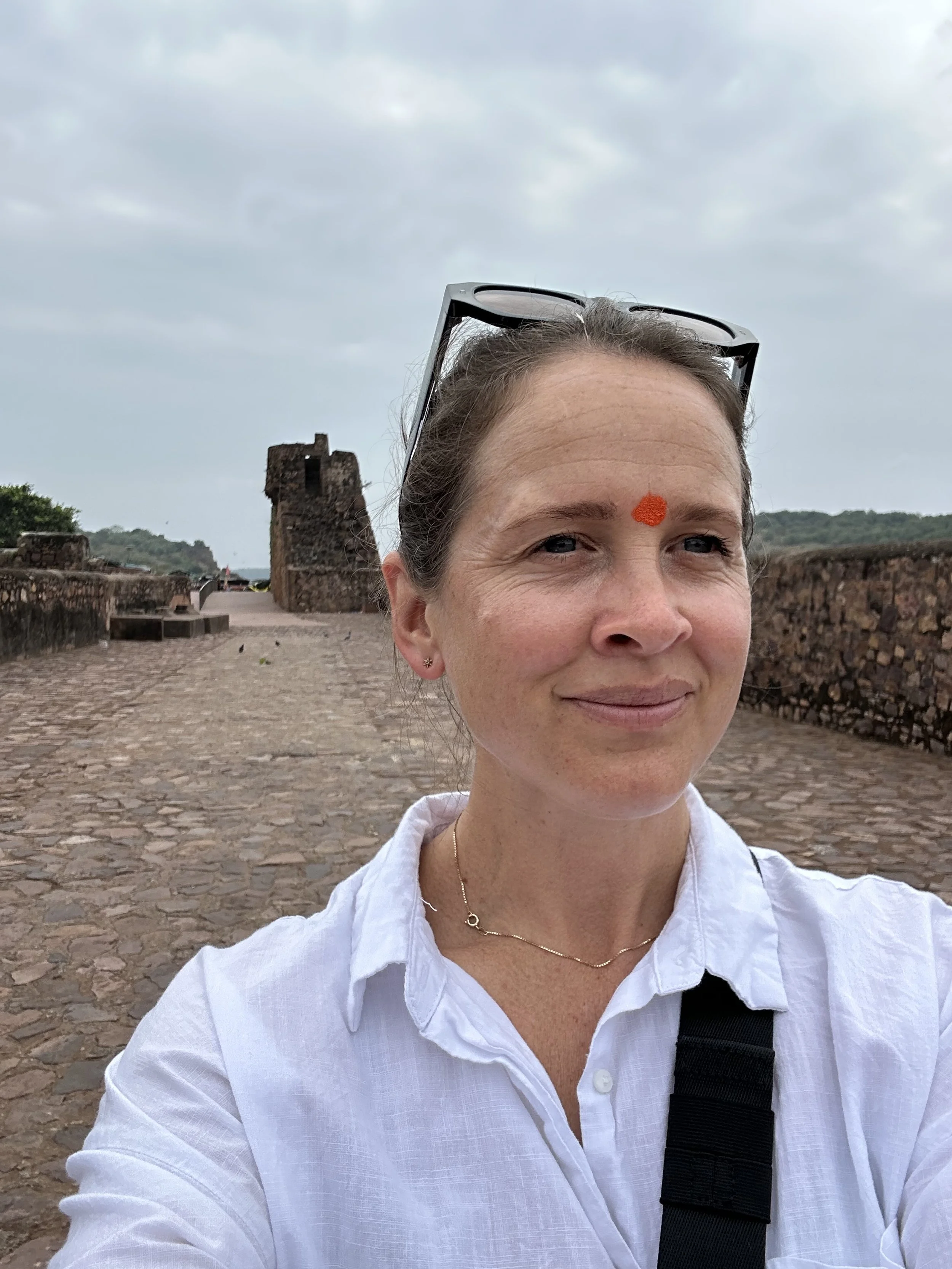 A woman with brown hair pulled back, wearing a patterned blouse with orange, pink, and blue designs, standing in front of an abstract, colorful painting with swirling shapes and pastel colors.