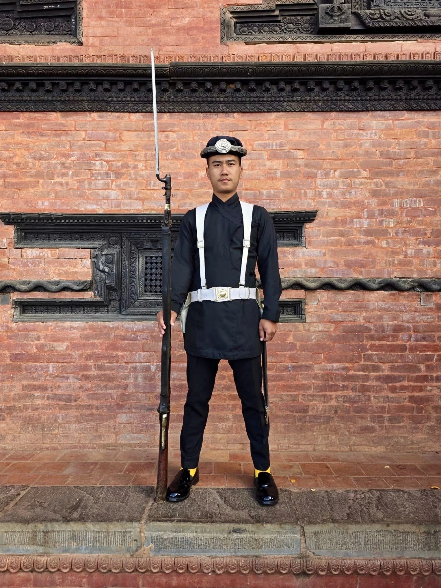 A young man dressed as a traditional British police officer, standing in front of a brick wall with ornate black wood carvings. He is holding a tall rifle and is wearing a dark uniform with a white belt and a cap with a badge.