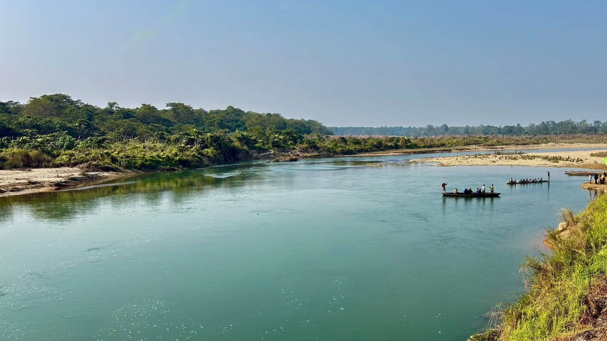 A river surrounded by greenery with two small boats carrying people on the water, and a few people standing on the riverbank.
