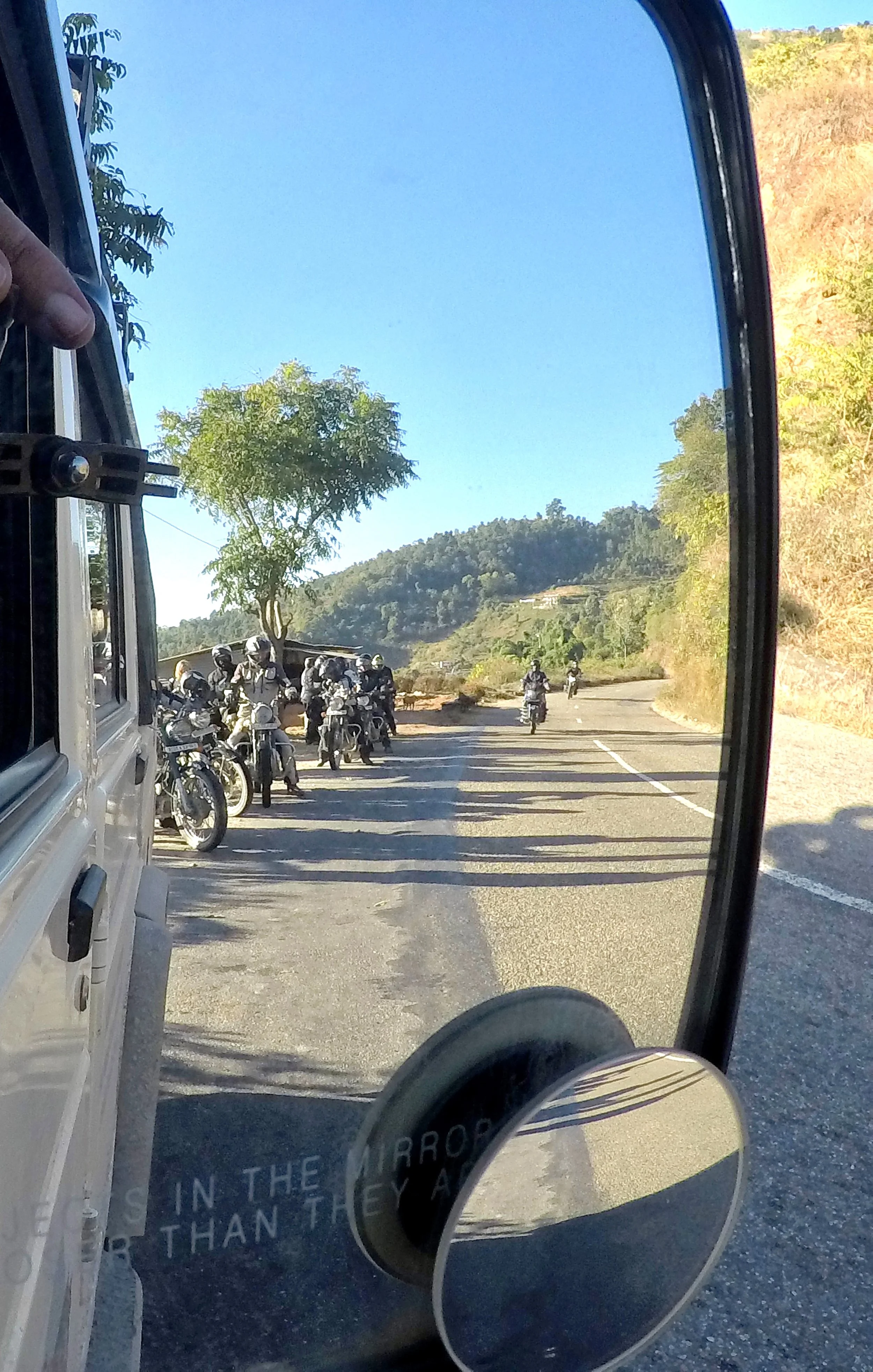 View through a vehicle mirror showing parked motorcycles on the roadside, a winding road, trees, hills, and clear blue sky.