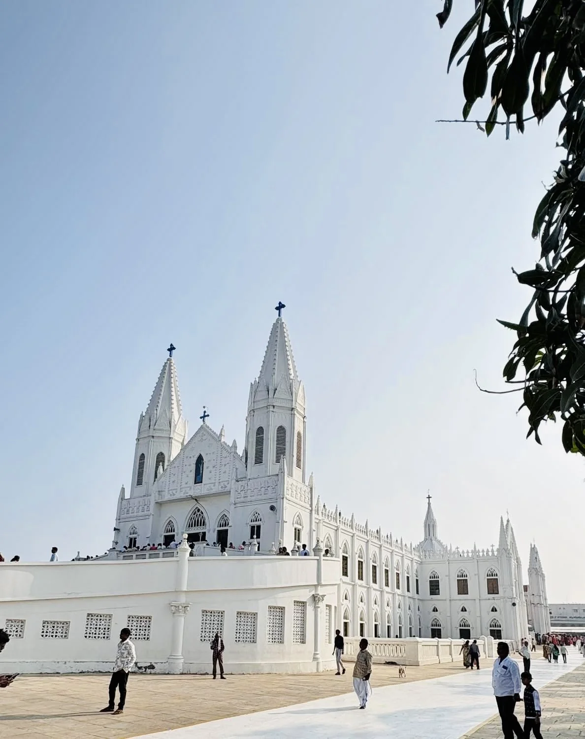 White church with twin steeples and crosses, people walking outside, clear sky, and green leafy branches in the top right corner.