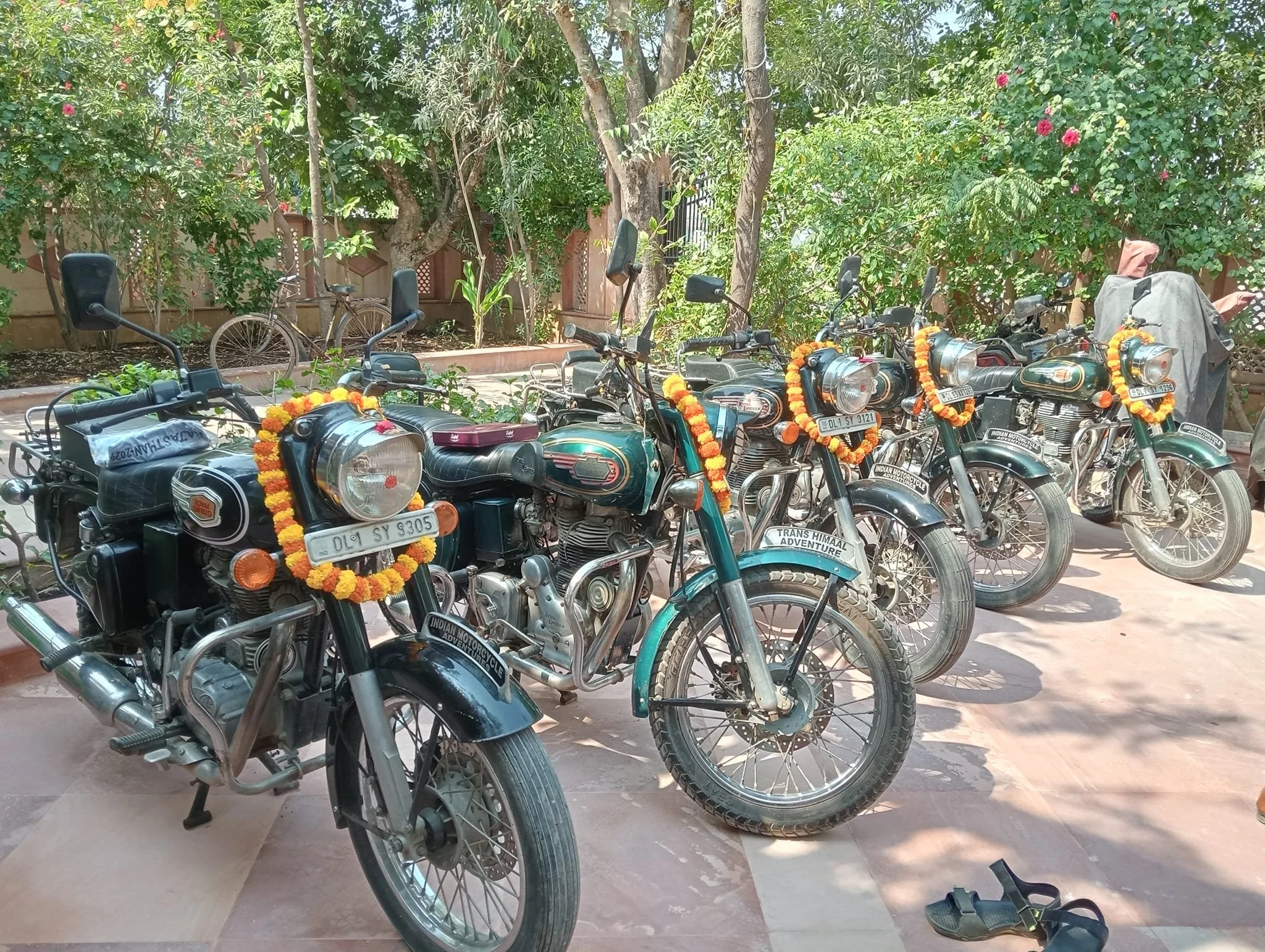 Six vintage motorcycles decorated with marigold garlands parked outdoors on a paved area with trees and plants in the background.
