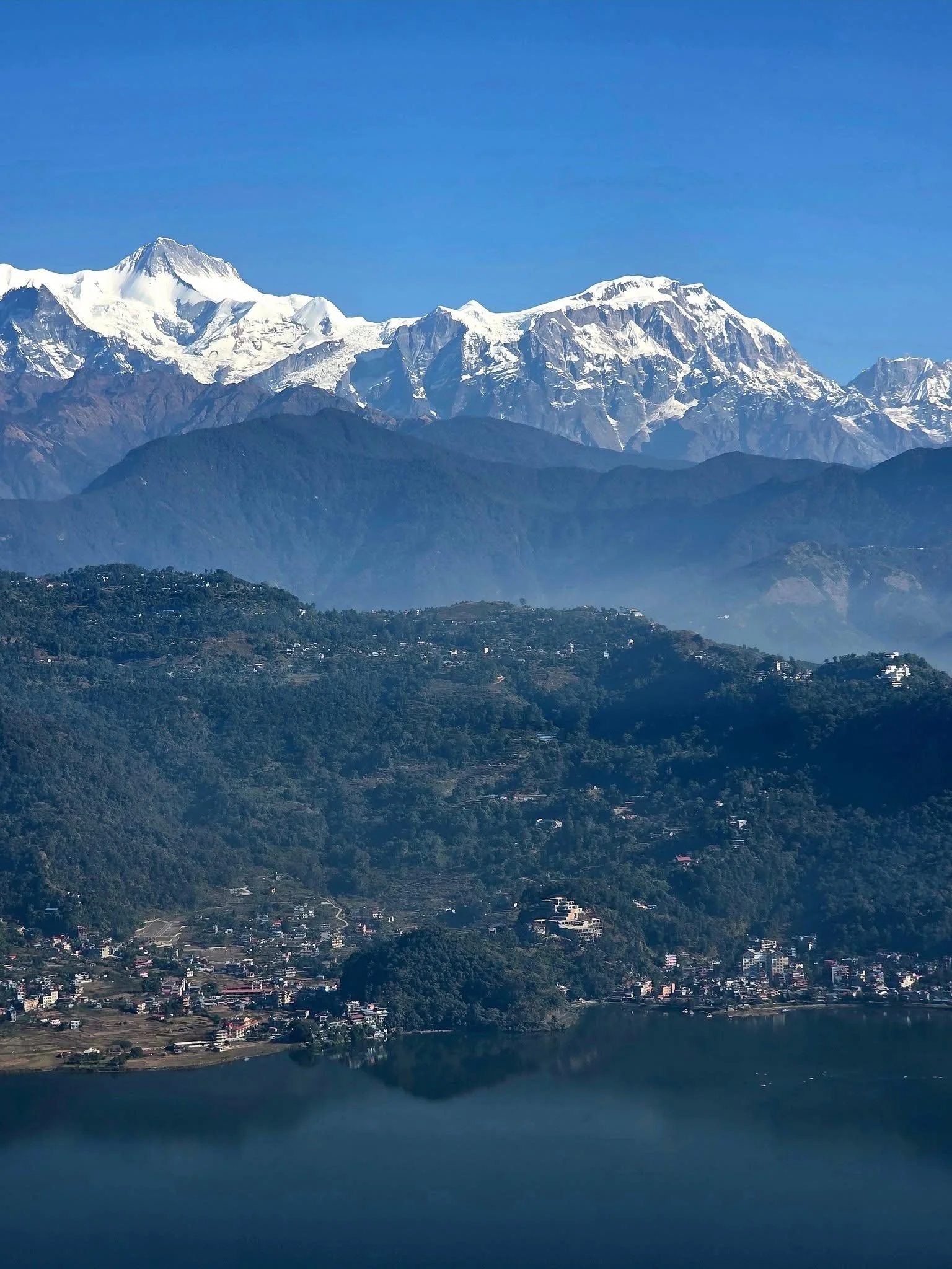 Snow-capped mountains over a lush green hillside and a calm lake in the foreground.