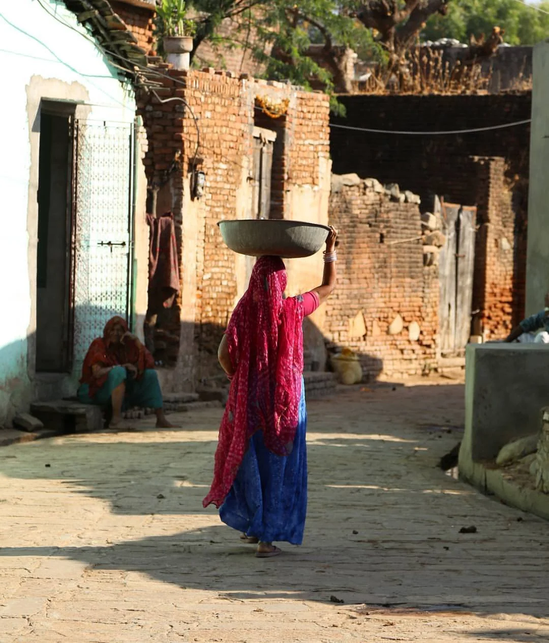 A woman dressed in colorful traditional attire carries a large bowl on her head while walking along a dusty street in a rural area with brick and concrete buildings and another woman sitting in the background.
