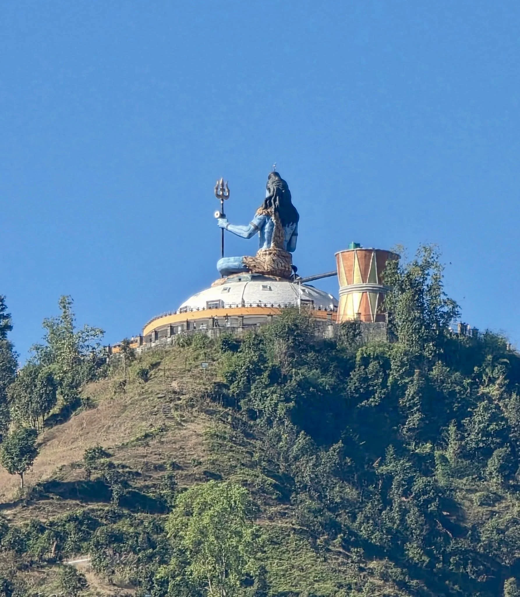 View of the giant statue of Lord Shiva on top of a hill, holding a trident in one hand, with greenery and trees around the hill and a clear blue sky in the background.