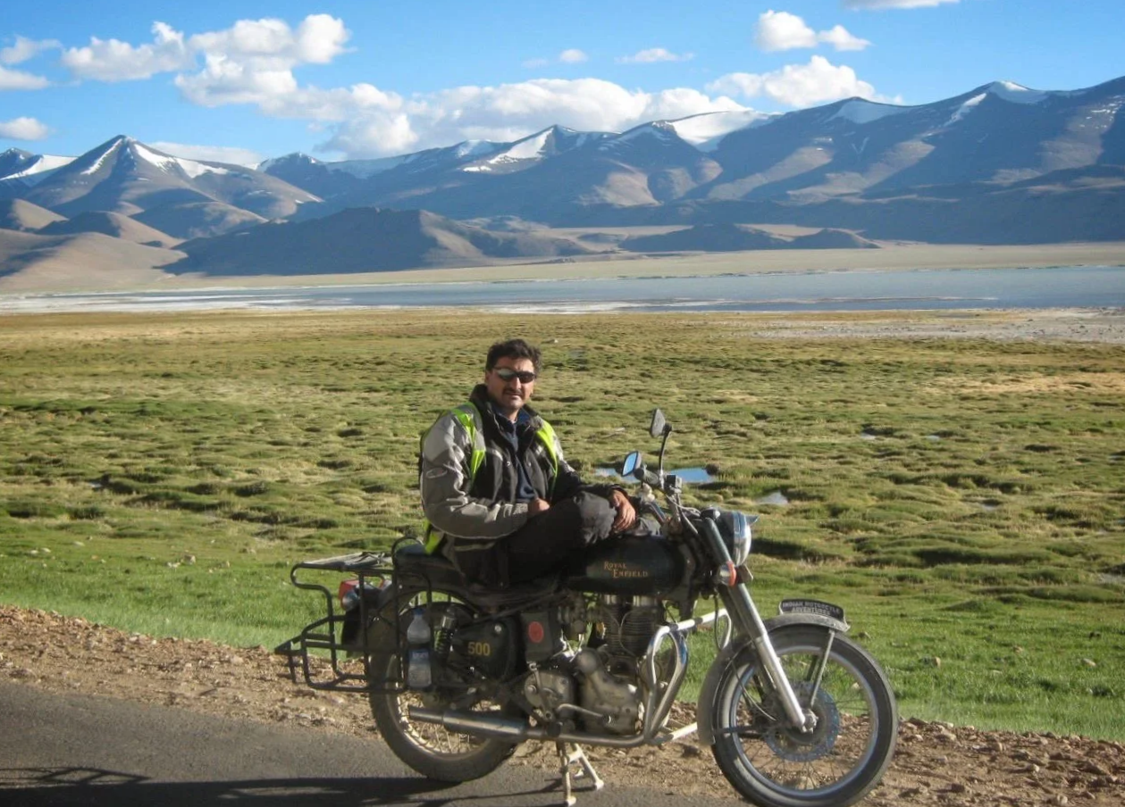 A man sitting on a black Royal Enfield motorcycle on a roadside in front of a grassy plain with mountains and snow-capped peaks in the background.