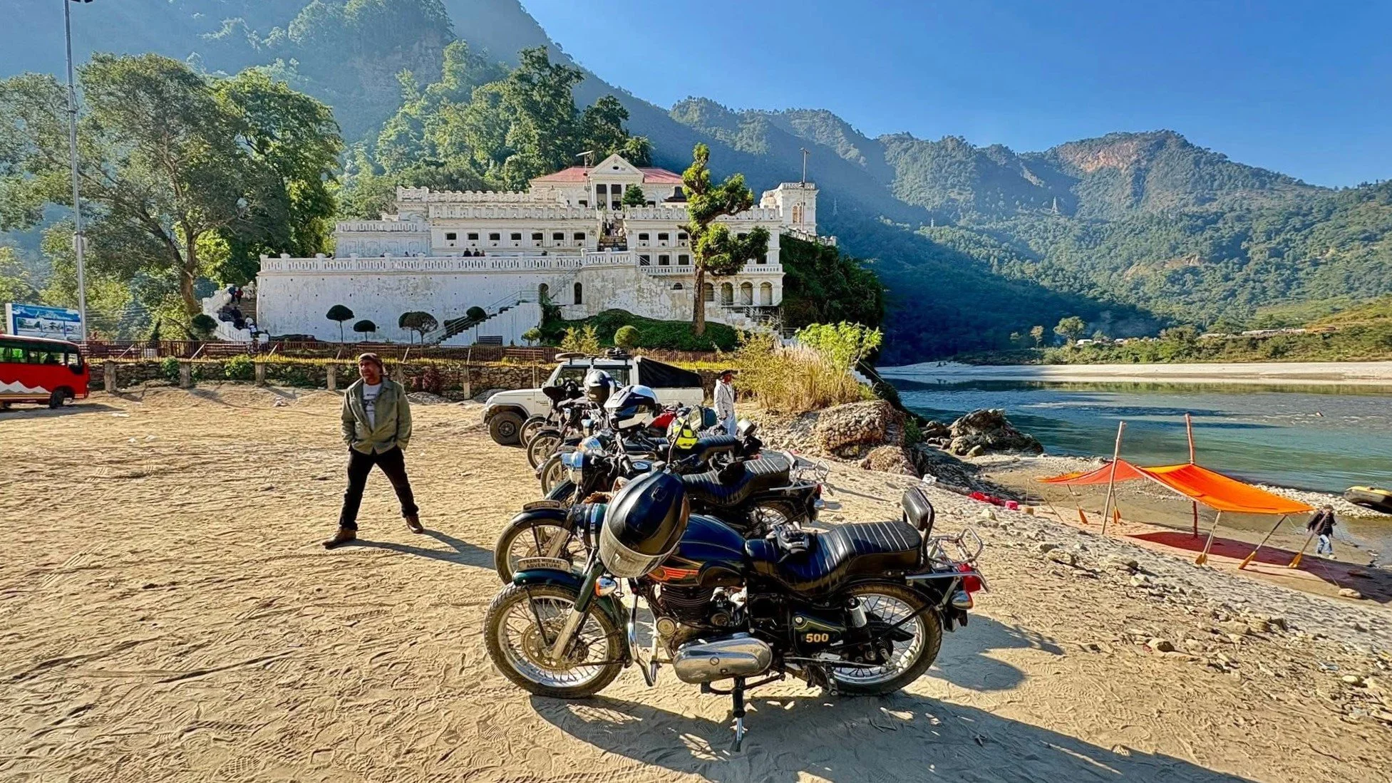 Motorcycles parked on a sandy riverside with a person standing nearby. A large white building is situated on a hill in the background with lush green mountains and a bright blue sky.