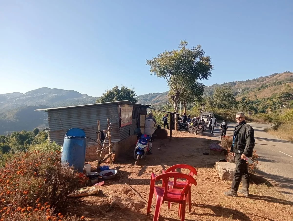A rural roadside scene with a small shack, motorcycles, a group of people, and a man in black standing near the road amid mountains and trees.
