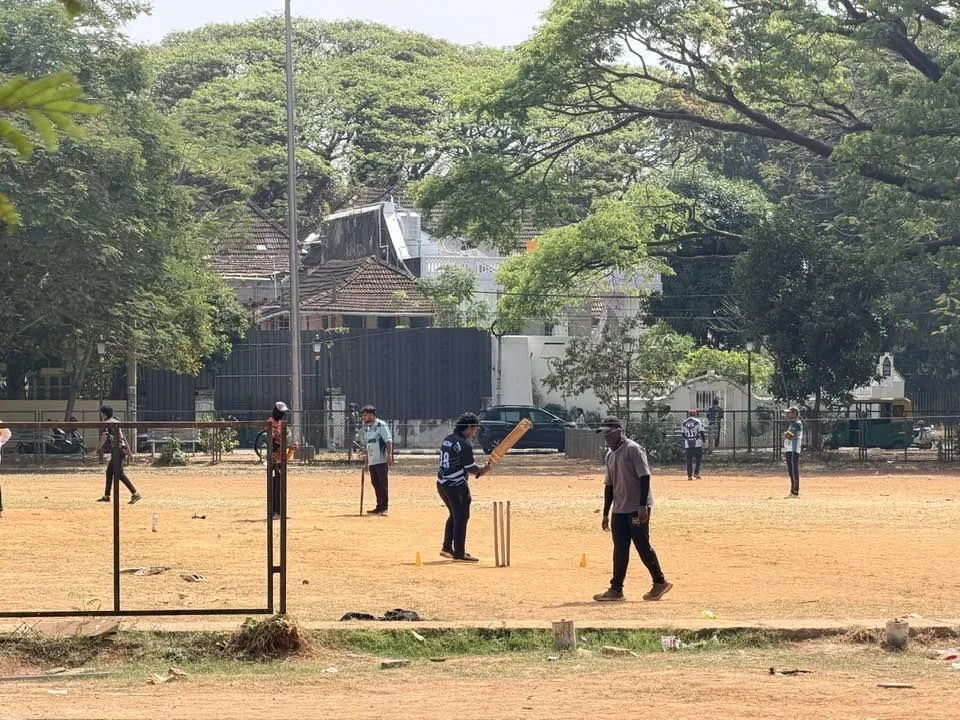People playing cricket on a dusty field with trees and buildings in the background.