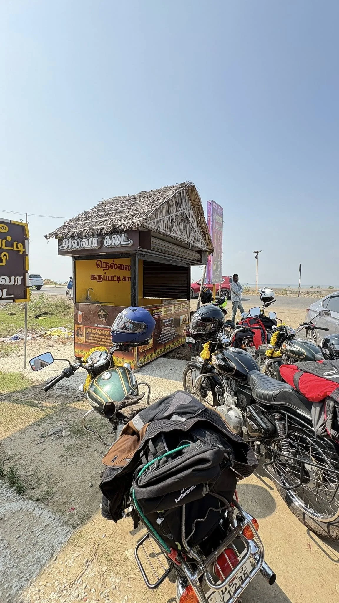A roadside food stall with a thatched roof in a sunny, open area. Several motorcycles are parked in front of the stall, some adorned with yellow garlands. In the background, there are a few people standing and some cars driving by.