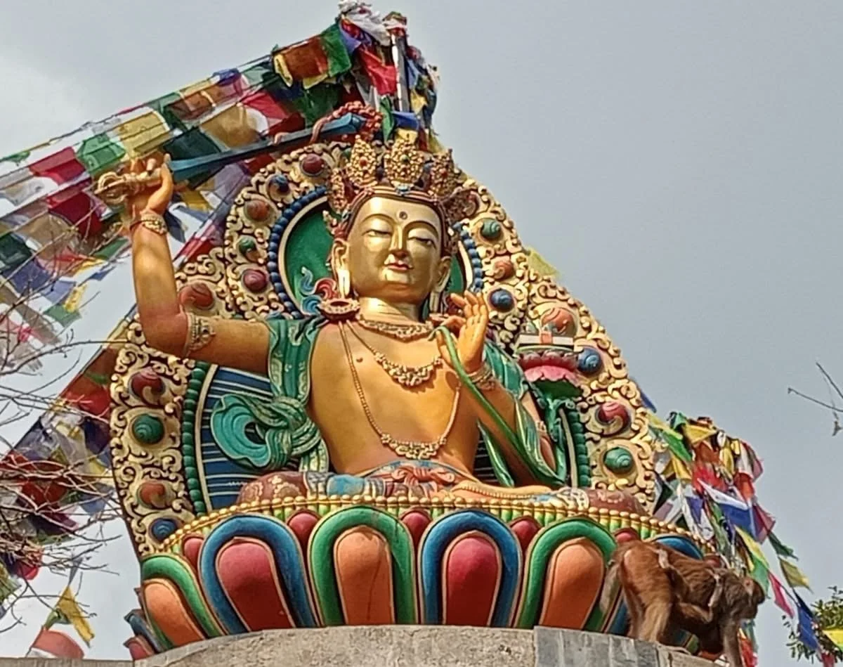 Colorful sculpture of a deity seated on a pedestal, adorned with jewelry and a crown, with multiple prayer flags in the background.