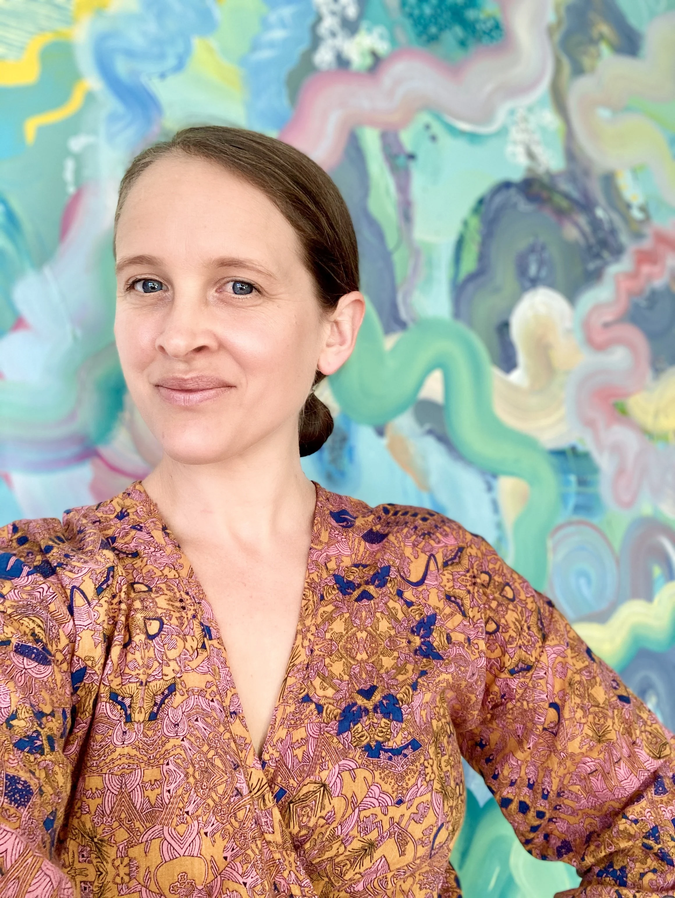 A woman with brown hair pulled back, wearing a patterned blouse with orange, pink, and blue designs, standing in front of an abstract, colorful painting with swirling shapes and pastel colors.