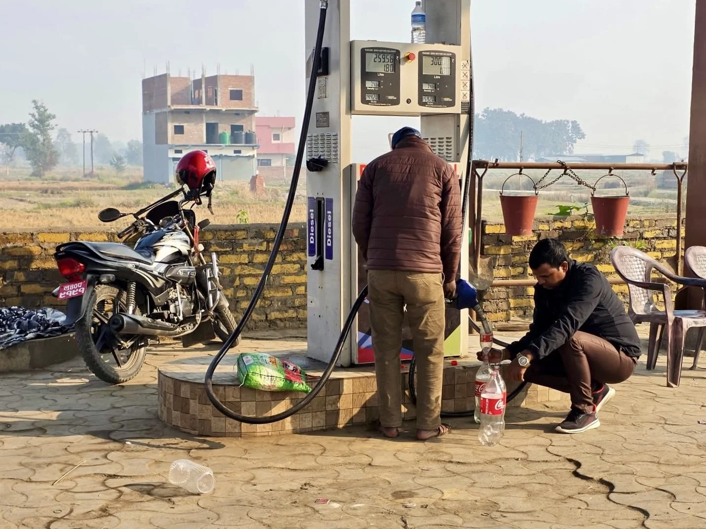 Two men filling a large plastic bottle with fuel at an outdoor fuel station, with a motorcycle parked nearby and a brick wall in the background.