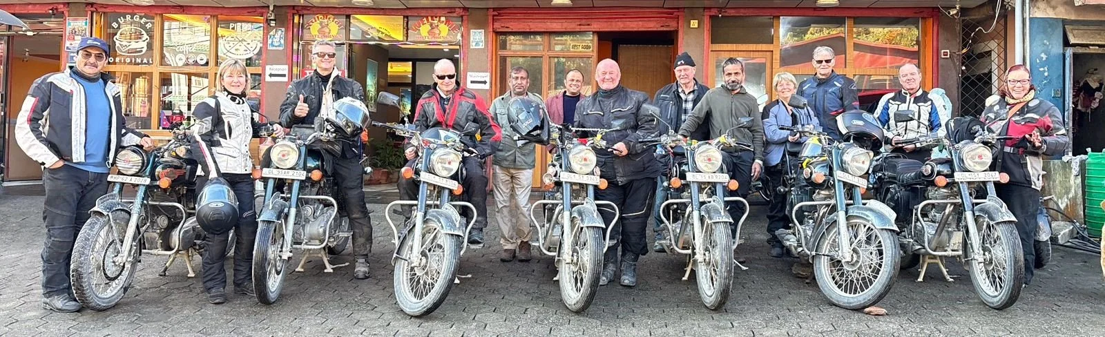 Group of twelve people standing behind motorcycle bikes outside of a restaurant or cafe.