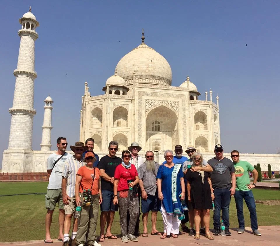 Group of people standing in front of the Taj Mahal, a white marble mausoleum with large domes and minarets in India, under a clear blue sky.