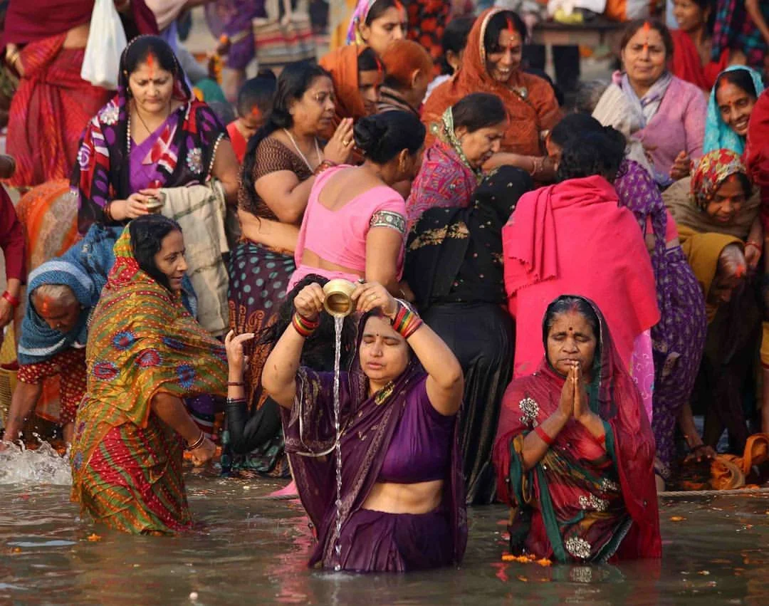 Women dressed in colorful traditional Indian attire participating in a religious ritual by a river, with some women praying and some standing in water.