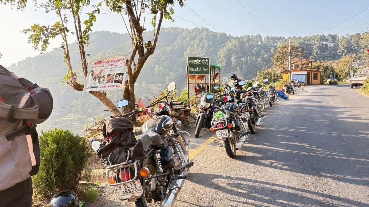 Line of parked motorcycles on the roadside near a green sign that reads 'Nagarkot View' in Nepali and English, with a tree, a small building, and hills in the background.