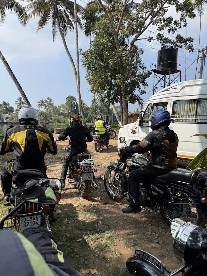 Group of motorcyclists riding along a dirt path lined with tall palm trees, with a white van parked nearby under large trees and a water tank on a metal structure in the background.
