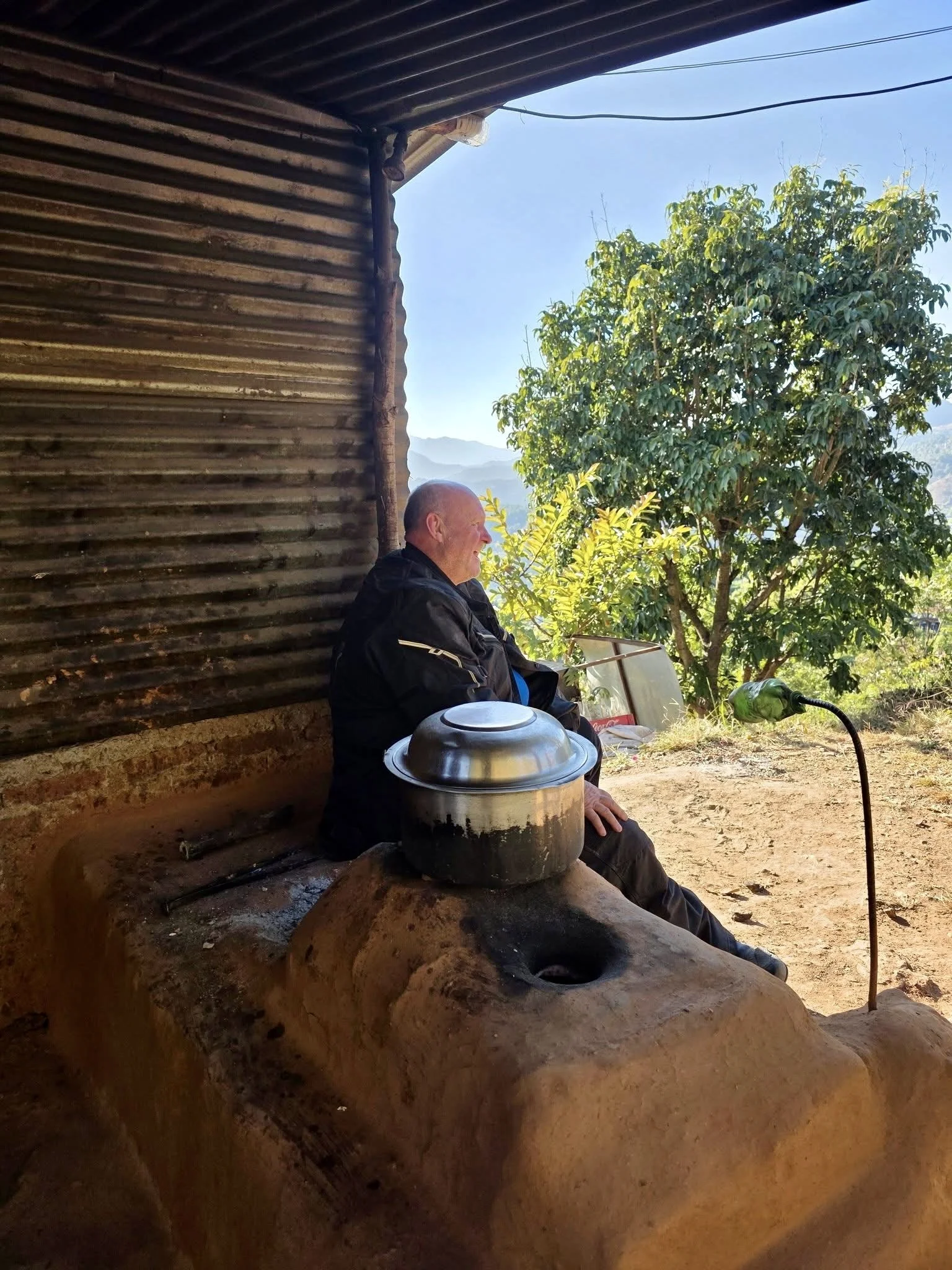 An older man sitting on a rustic outdoor stove under a partially covered area, with a large cooking pot in front of him. He is outdoors next to a tree with green foliage, on a dirt ground, and there is a bright blue sky in the background.
