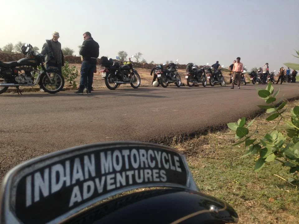 Motorcycles parked on the side of a road with people standing nearby, and a motorbike helmet with a sticker that reads 'Indian Motorcycle Adventures' in the foreground.