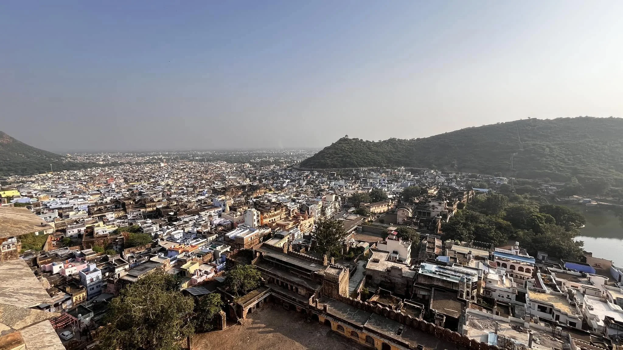 A view of a densely populated city with numerous small buildings, surrounded by hills and a body of water on the right side under a clear sky.