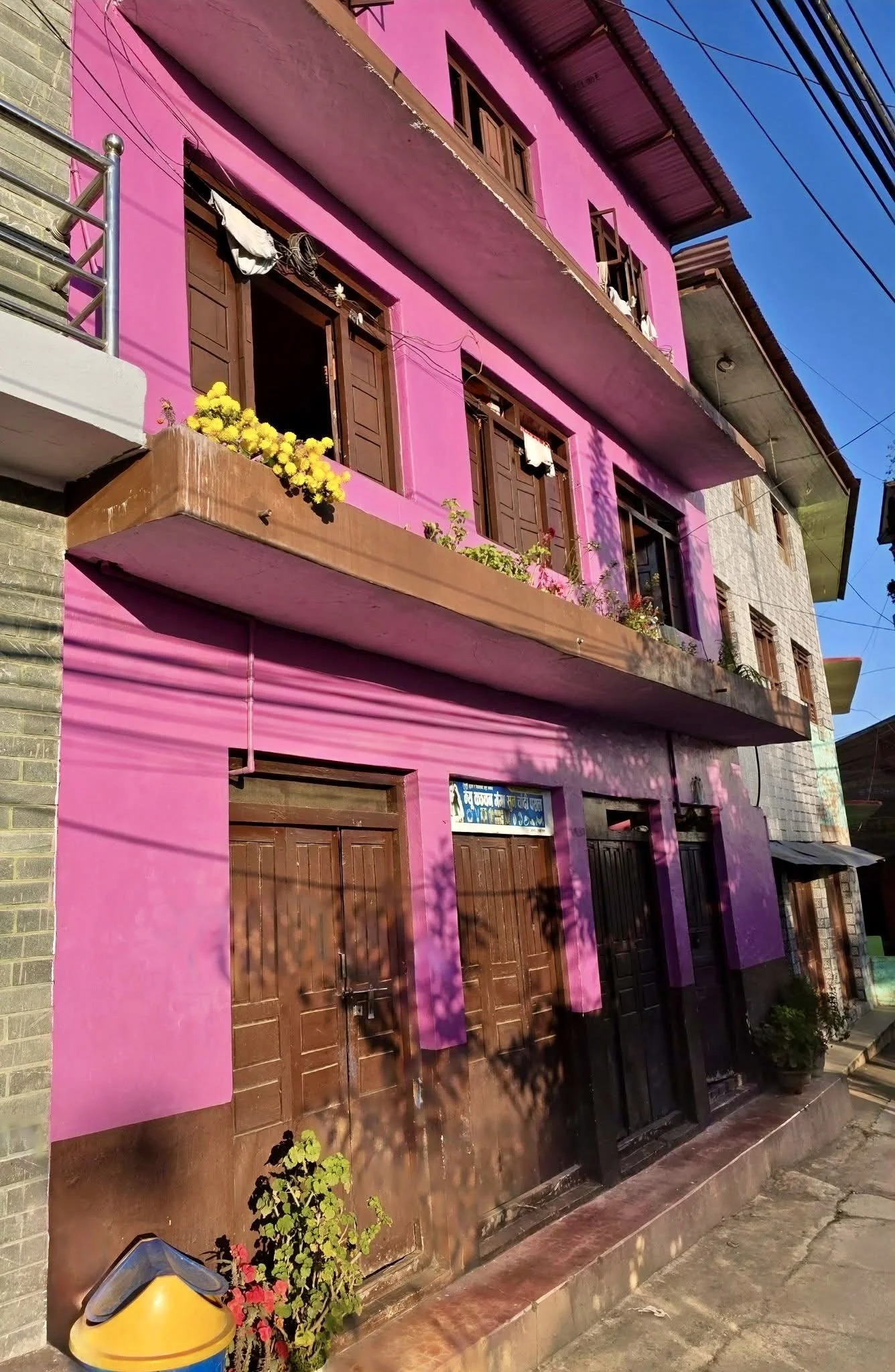 A three-story pink building with brown wooden doors and windows, flower pots on the balconies, and shadows of trees cast on the facade, located on a sunny street.